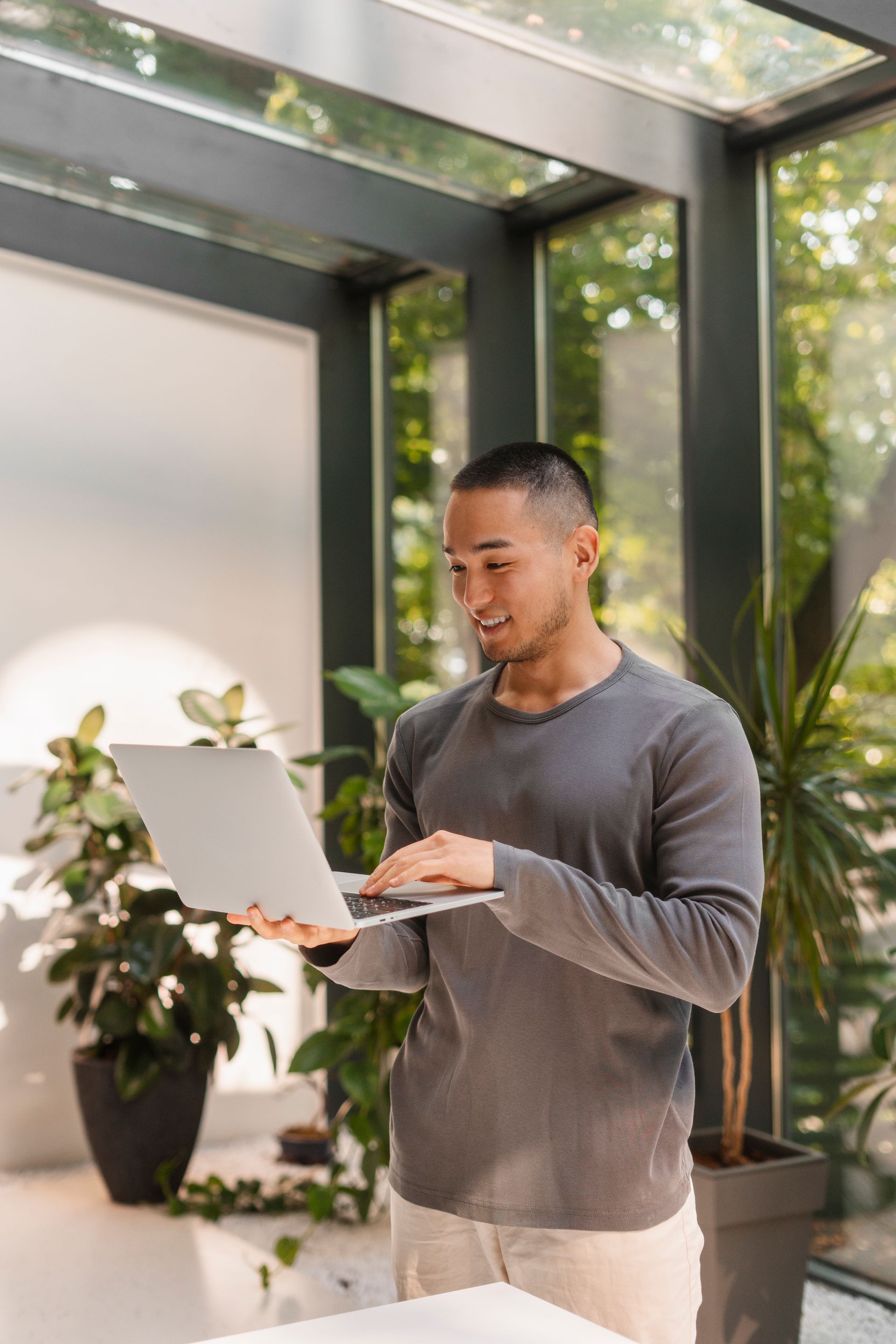 Person using a laptop in a bright indoor garden space with large windows and potted plants