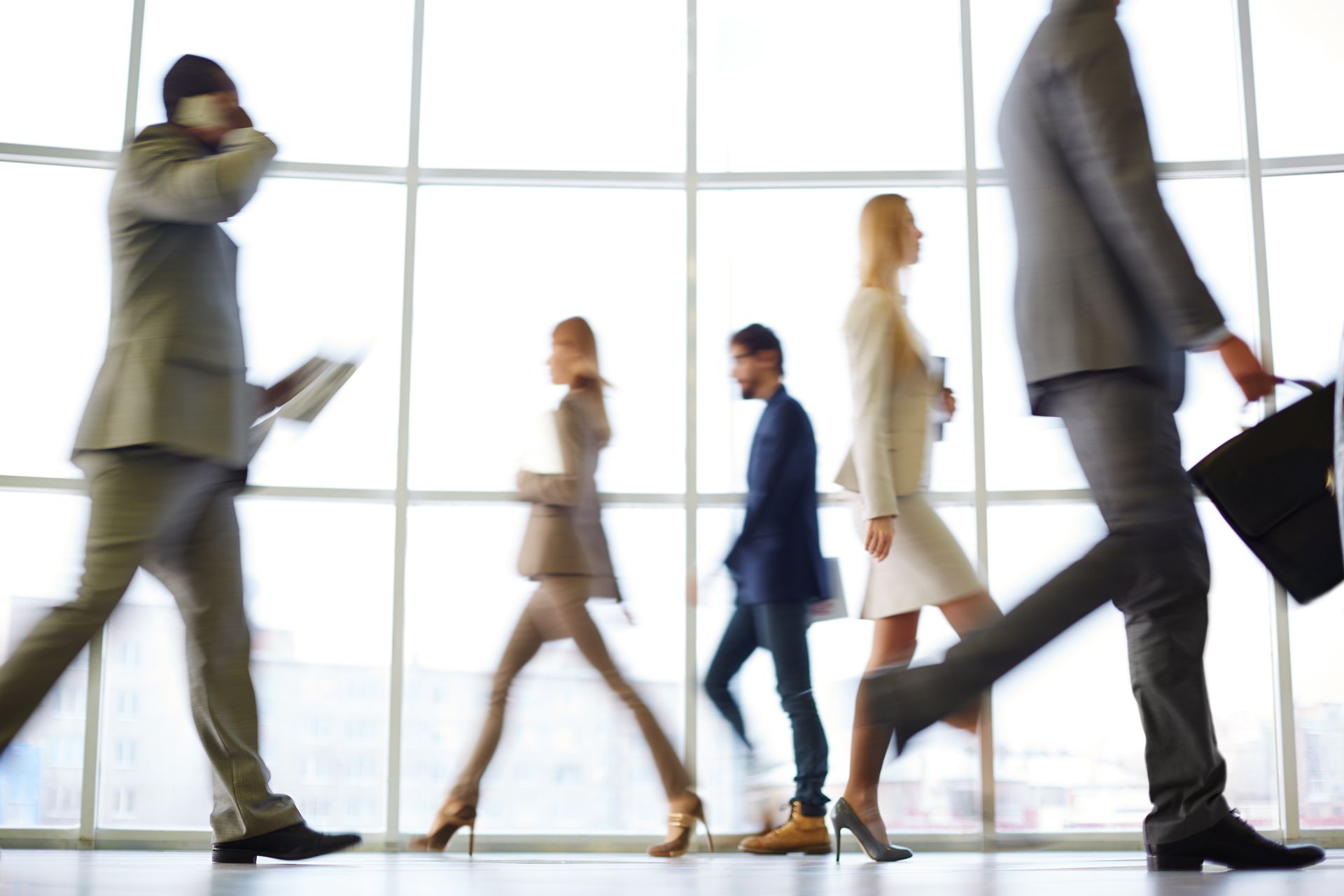 Professionals in business attire walking quickly past large floor-to-ceiling windows in a bright office hallway.