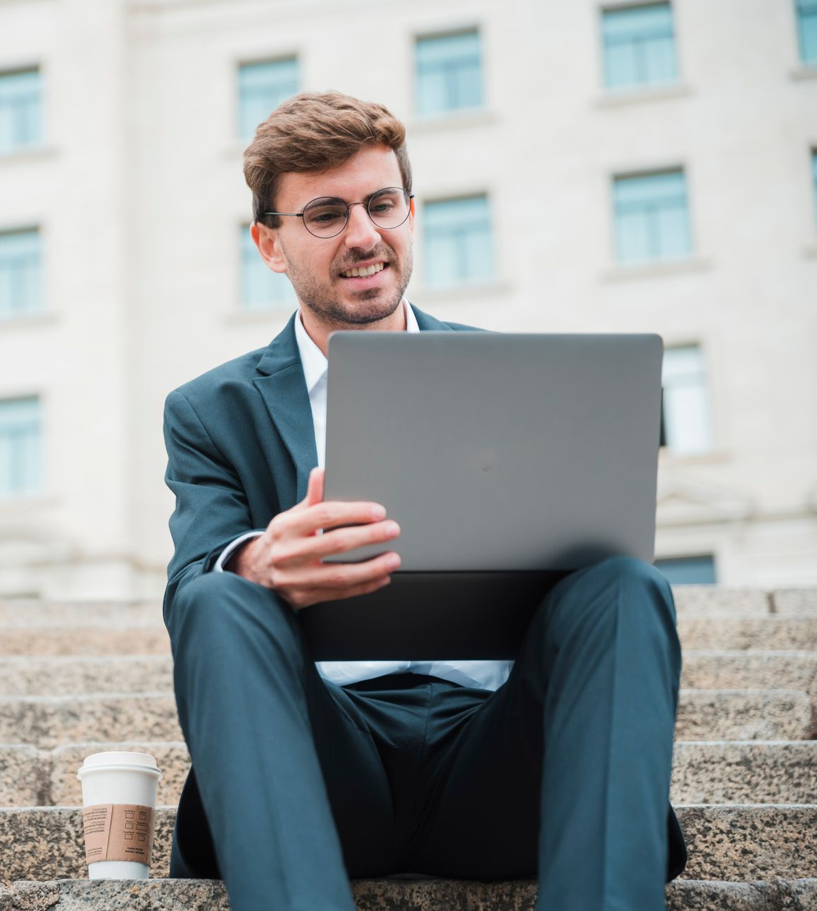 A person in a business suit sitting on stone steps outdoors, smiling while looking at a laptop with a coffee cup nearby.