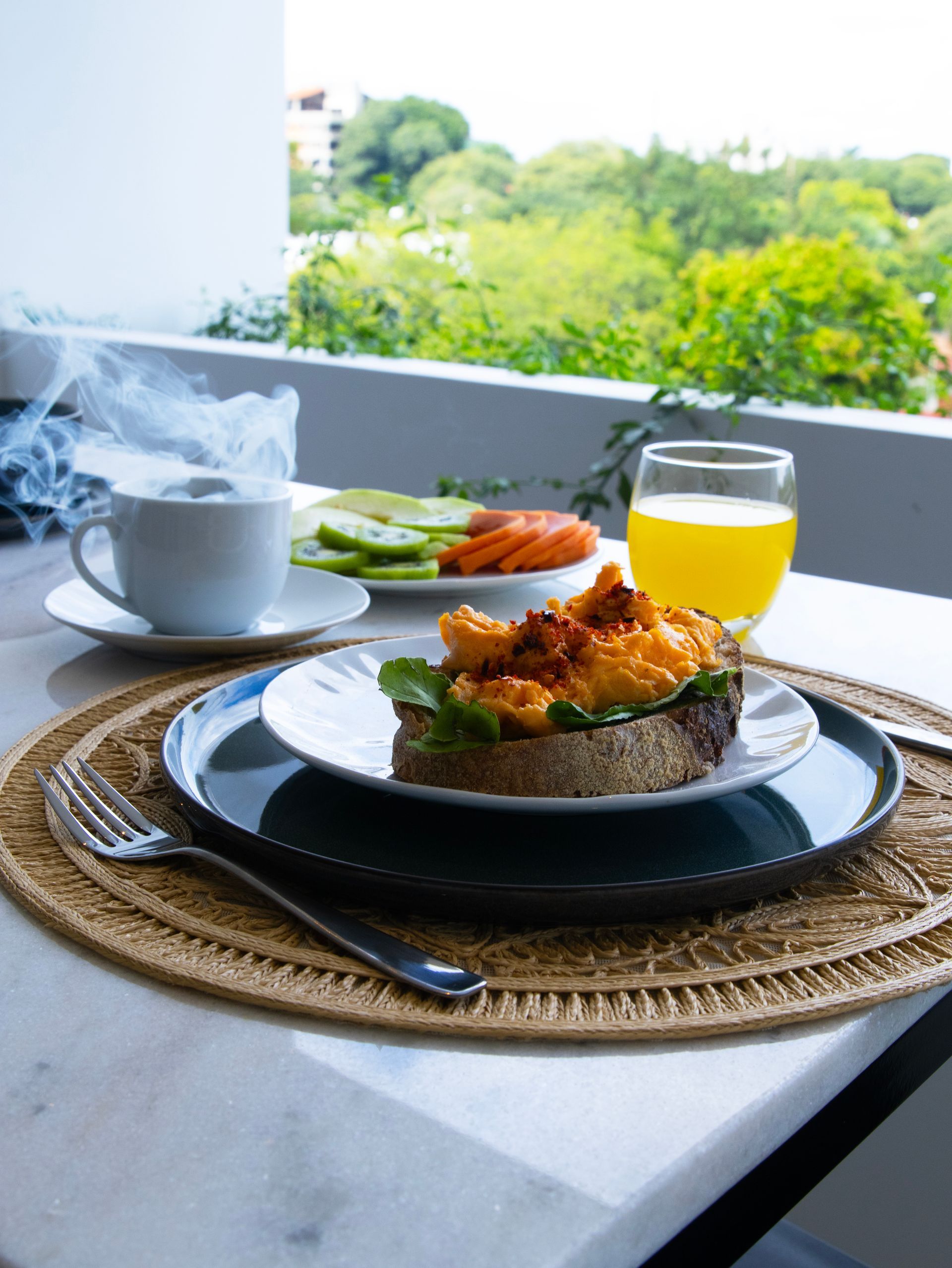 Desayuno en un balcón: huevos revueltos sobre tostadas, café, jugo y verduras en rodajas, con vista verde.
