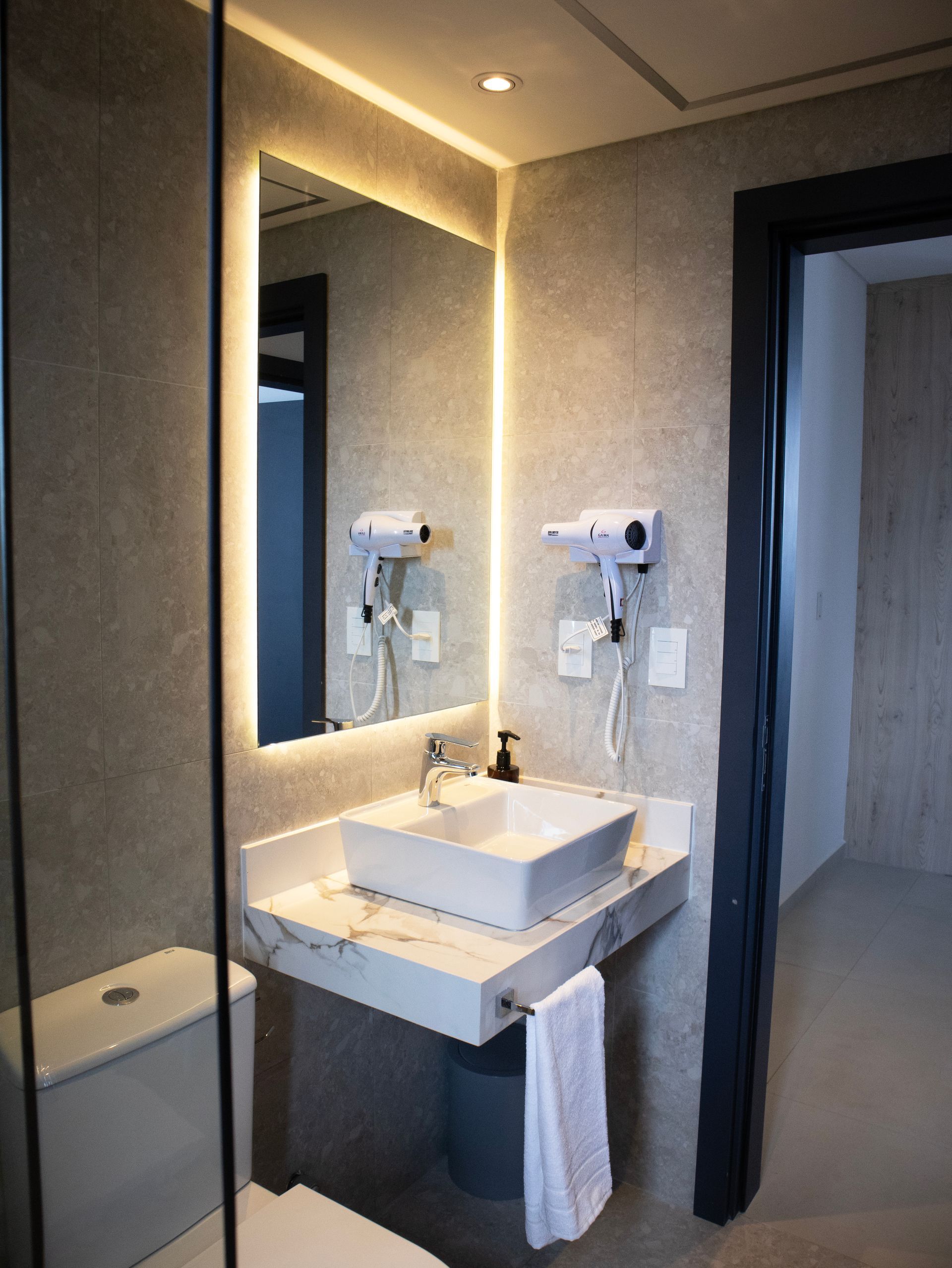 Baño con espejo iluminado encima del lavabo y secador de pelo montado en la pared.