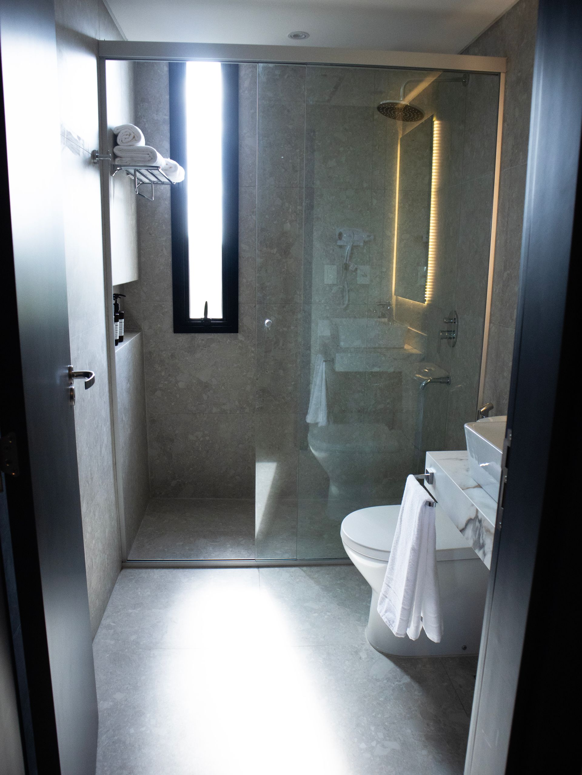 Pequeño baño moderno con ducha con mampara de cristal, inodoro y lavabo. Paredes y suelo grises.