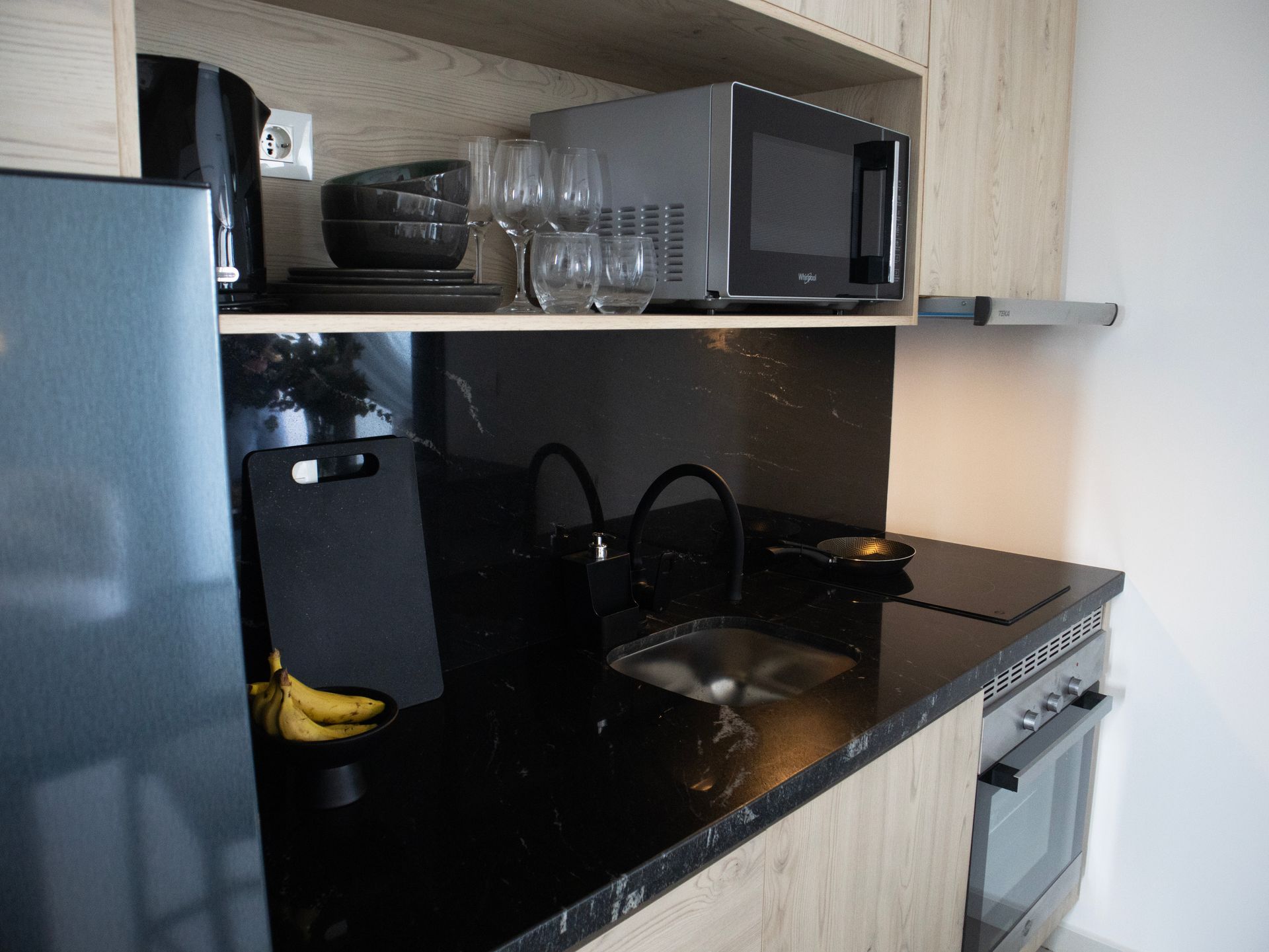Cocina pequeña con encimeras negras, microondas, fregadero y gabinetes abiertos.