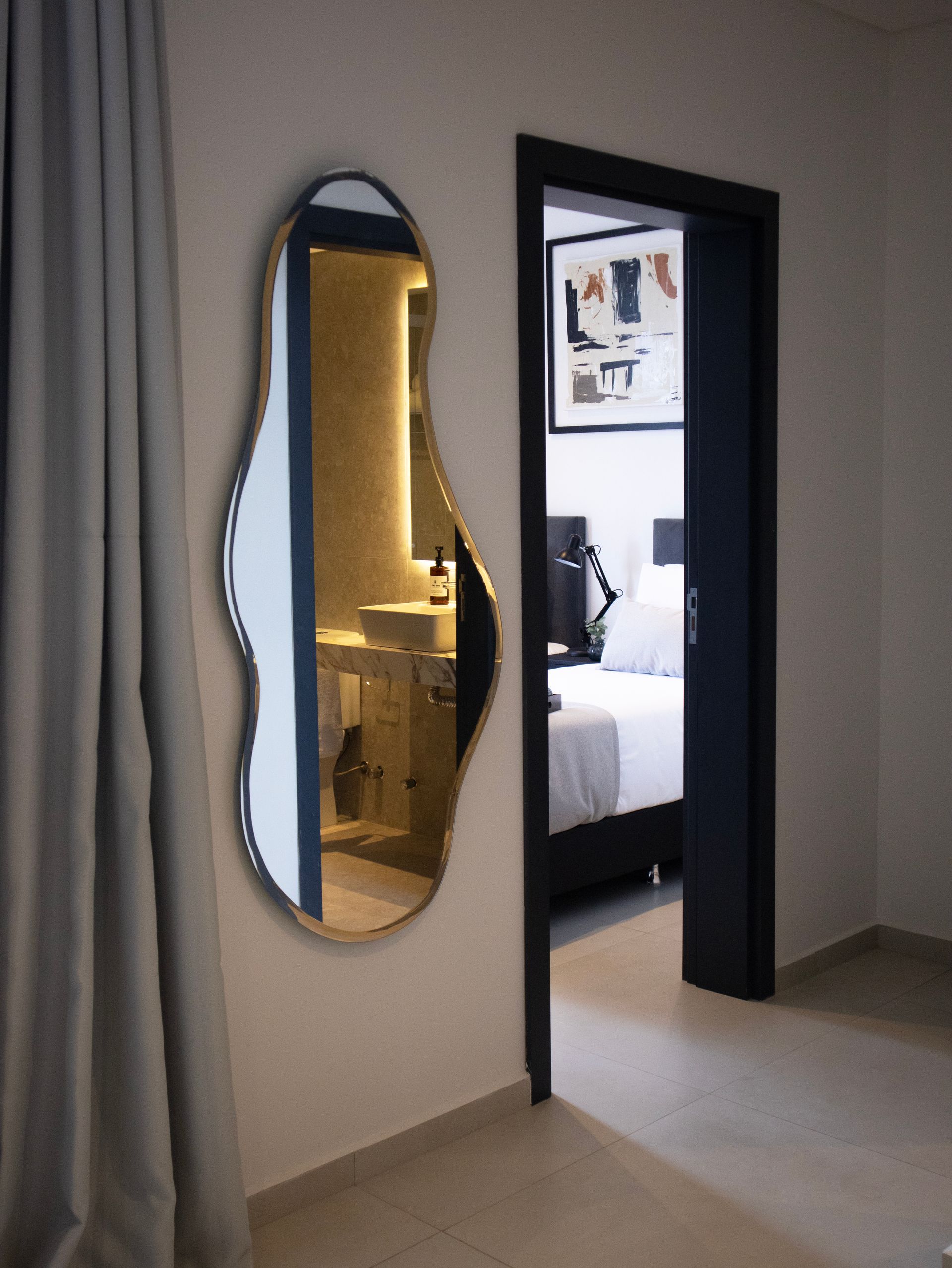 Espejo de forma irregular en una pared, que refleja un baño y una puerta que conduce a un dormitorio.