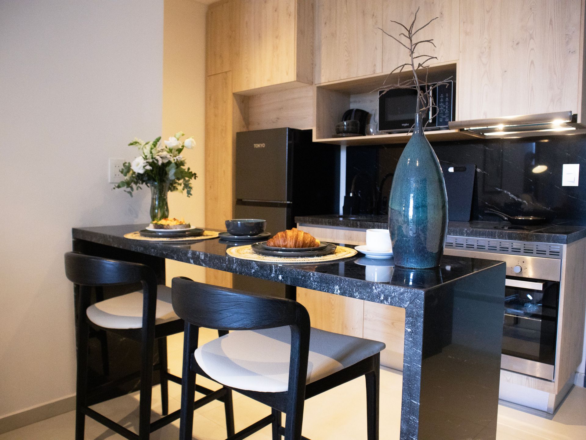Cocina moderna con encimera negra y dos taburetes. Incluye refrigerador, microondas y jarrón negros.