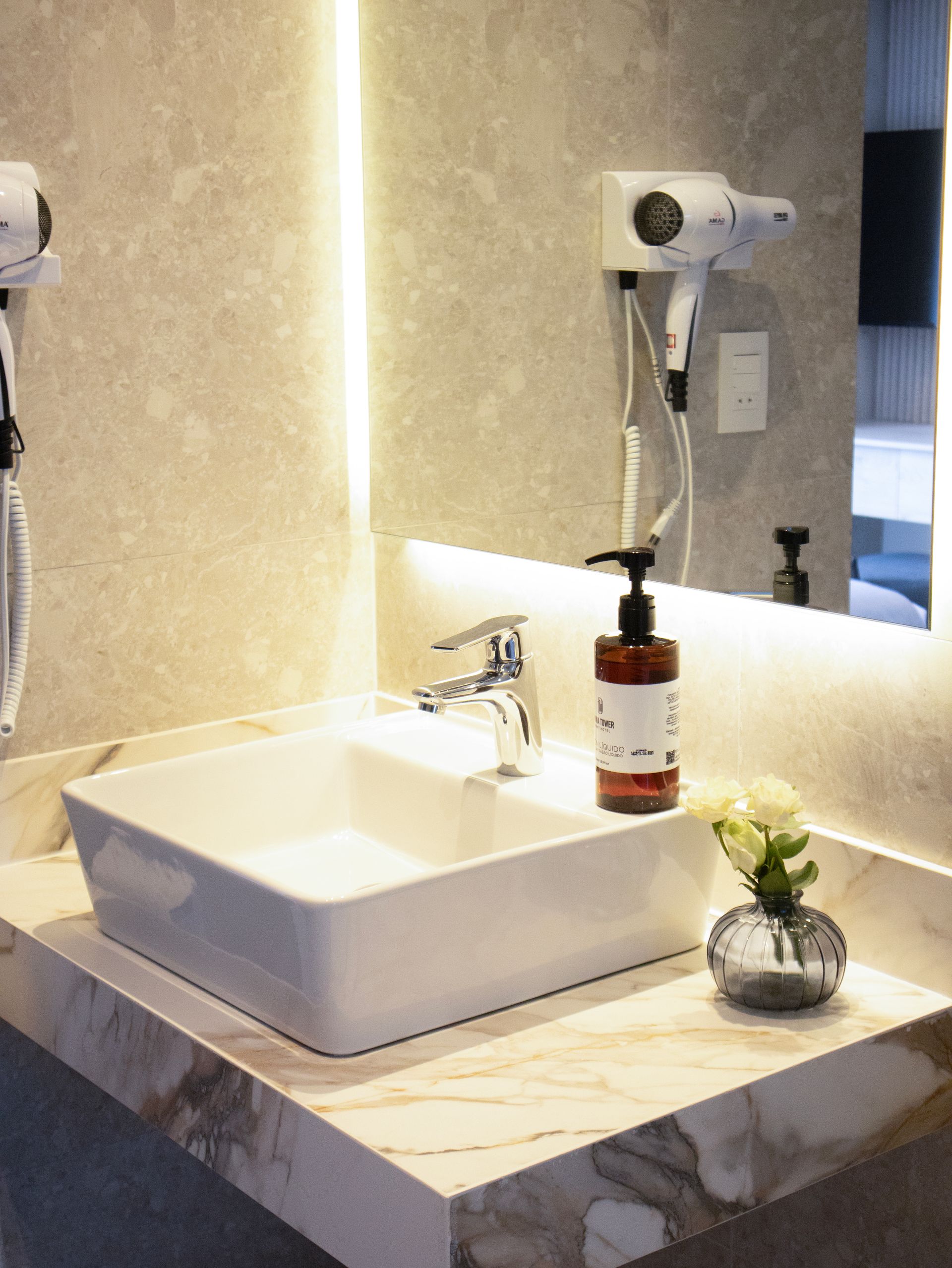 Lavabo de baño con encimera de mármol, un dosificador de jabón y un pequeño jarrón con flores.