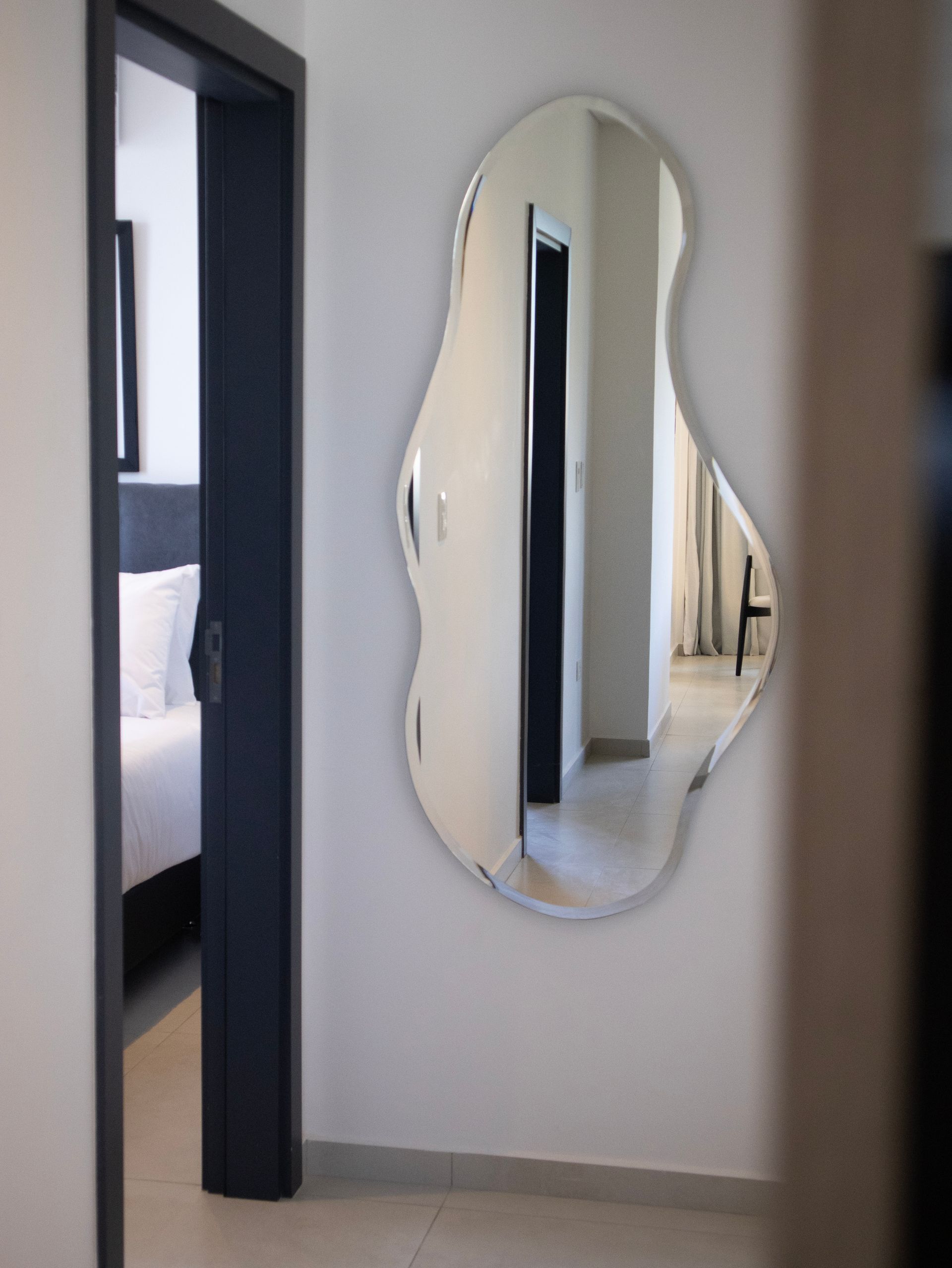 Un espejo de forma abstracta sobre una pared blanca refleja la puerta de un dormitorio y un pasillo.