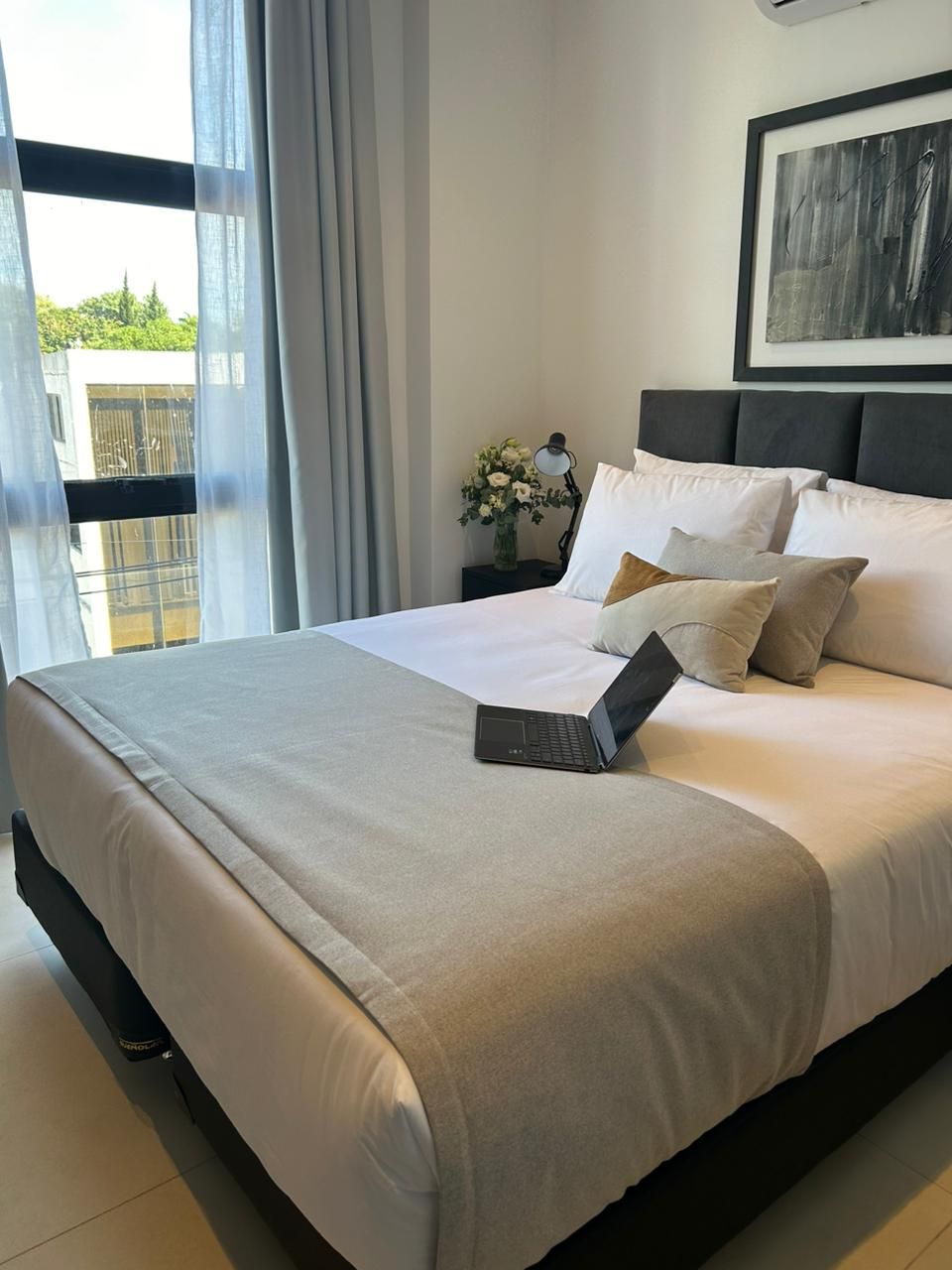Dormitorio con cama, almohadas, computadora portátil, ventana con cortinas transparentes y obras de arte.
