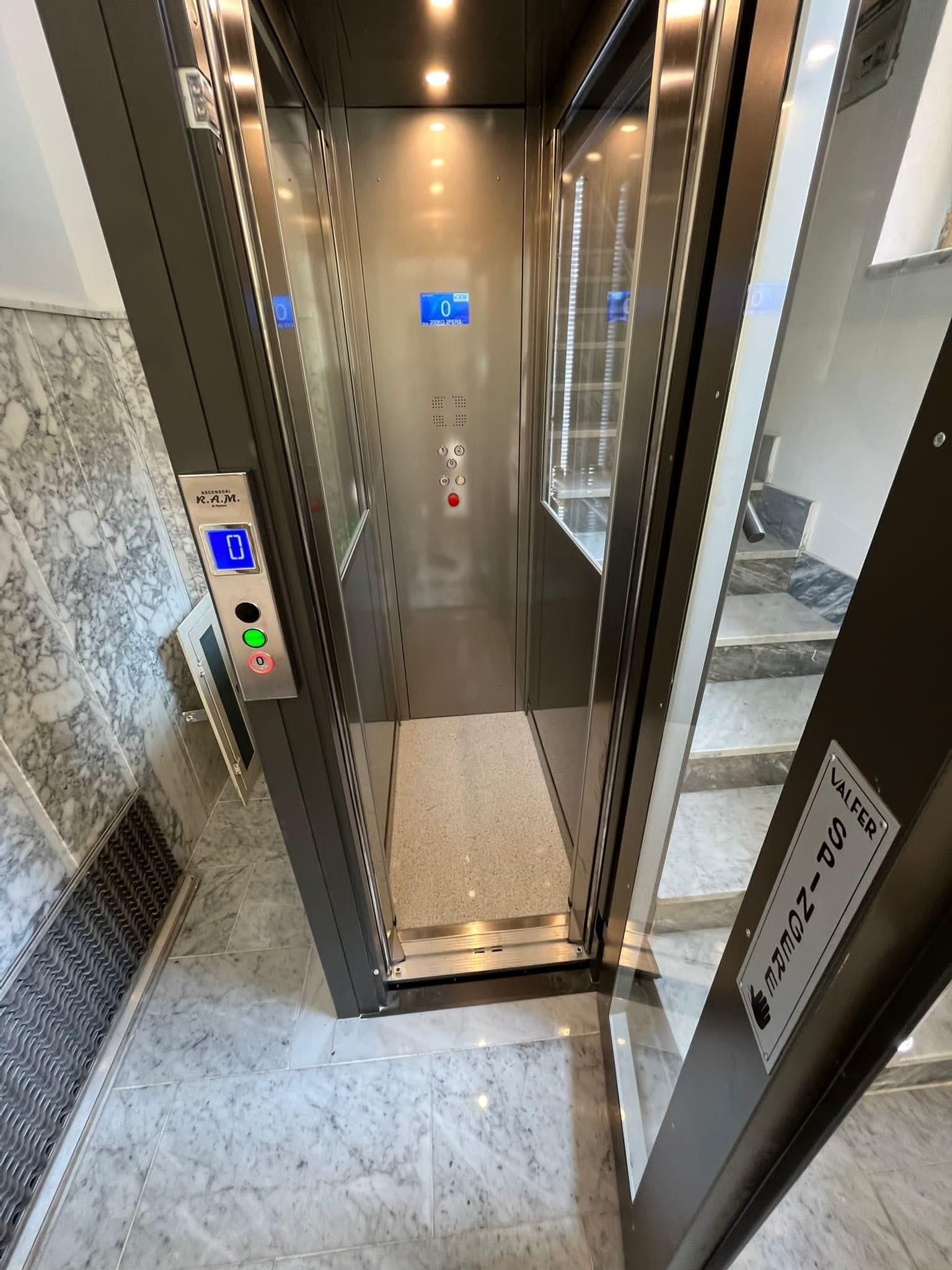 Ascensore in un edificio, aperto. Interni in acciaio inox, porte in vetro, con pulsanti di accesso sulla sinistra.