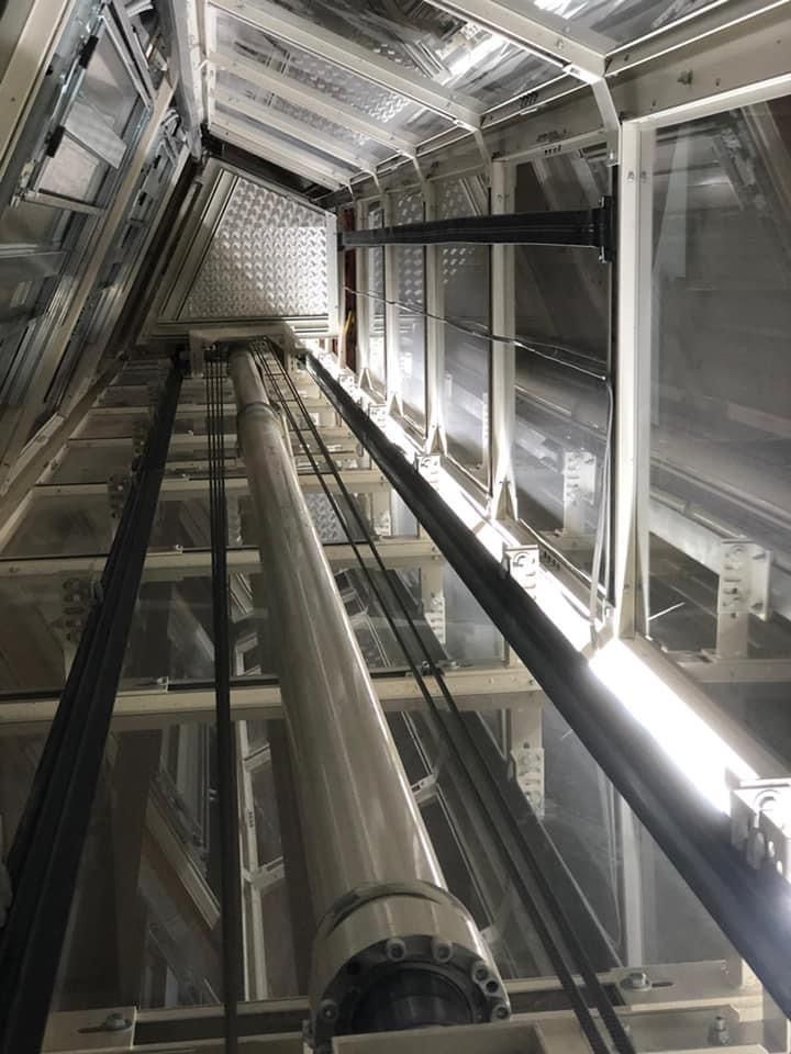 Interno del vano ascensore, vista dall'alto. Cilindro idraulico argentato e cavi che corrono verticalmente. Architettura in acciaio e vetro.