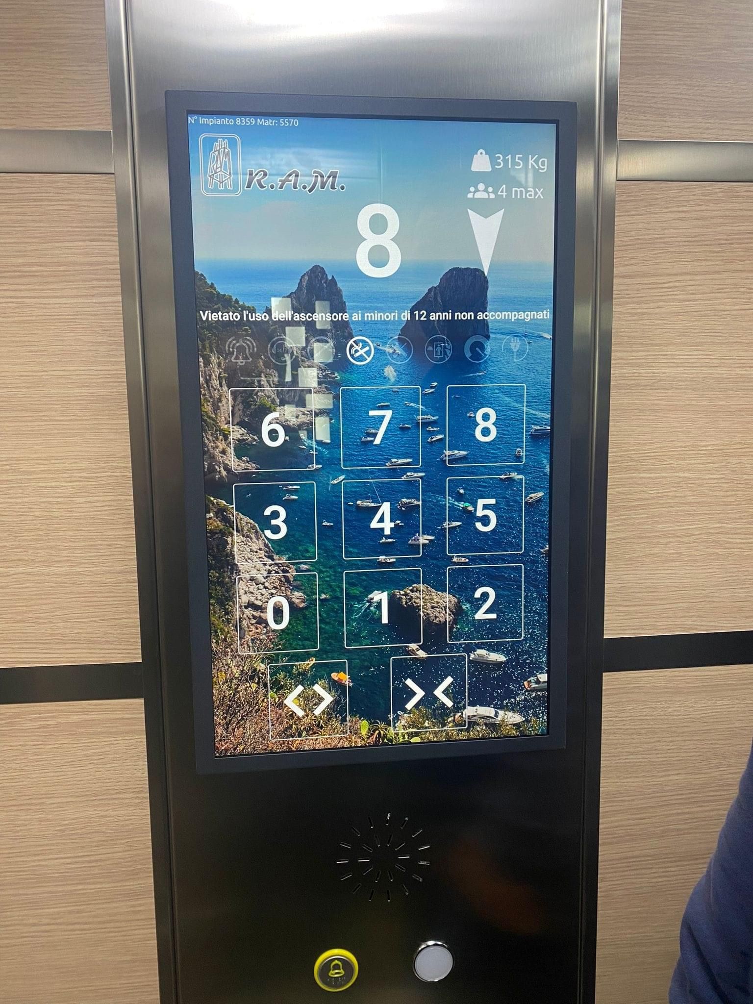 Pannello di controllo dell'ascensore con touch screen che mostra i numeri dei piani e uno sfondo panoramico. Il piano attuale è l'8.