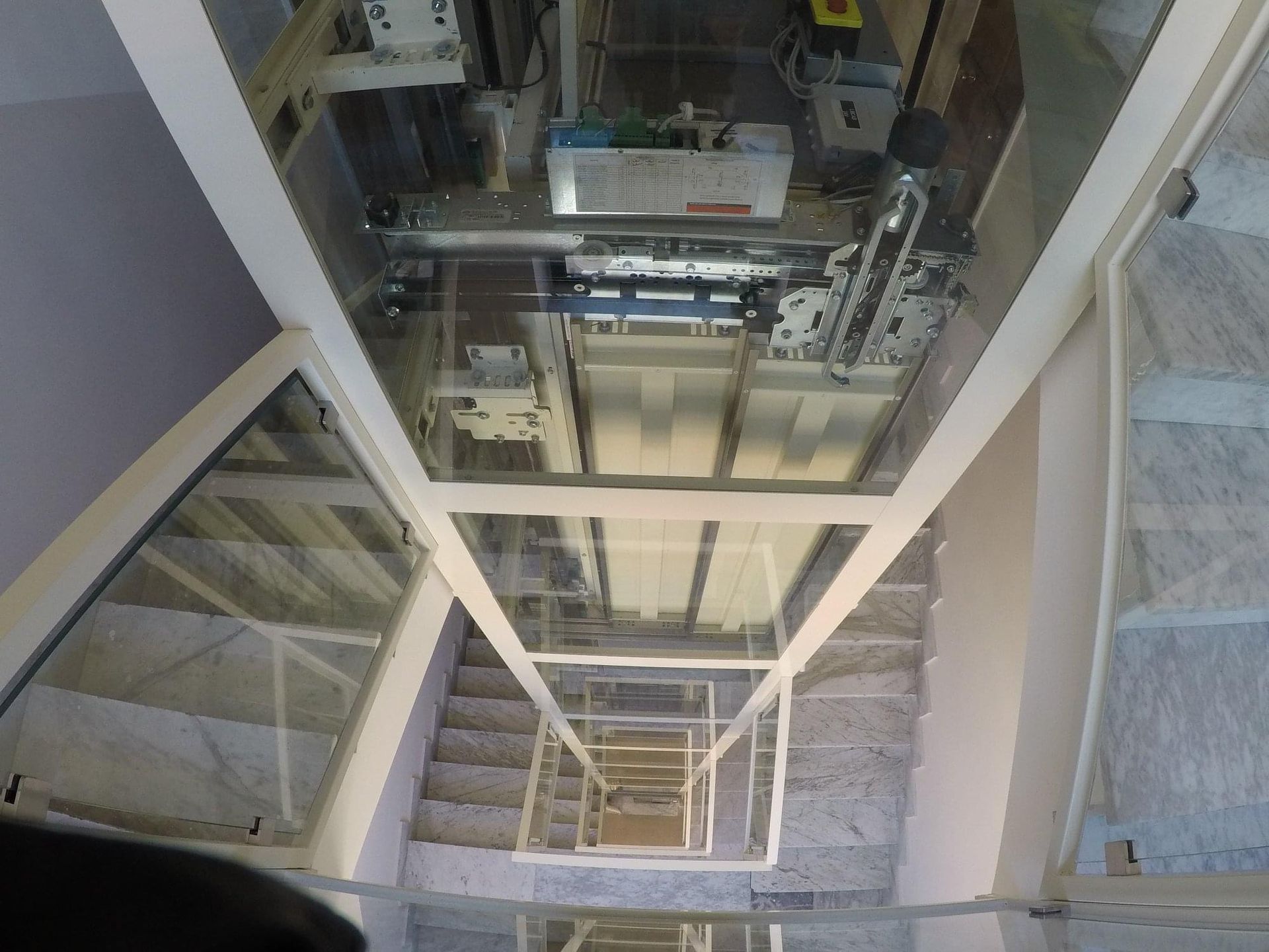 Guardando lungo il vano ascensore in vetro, si vedono macchinari, scale e un soffitto visibile.