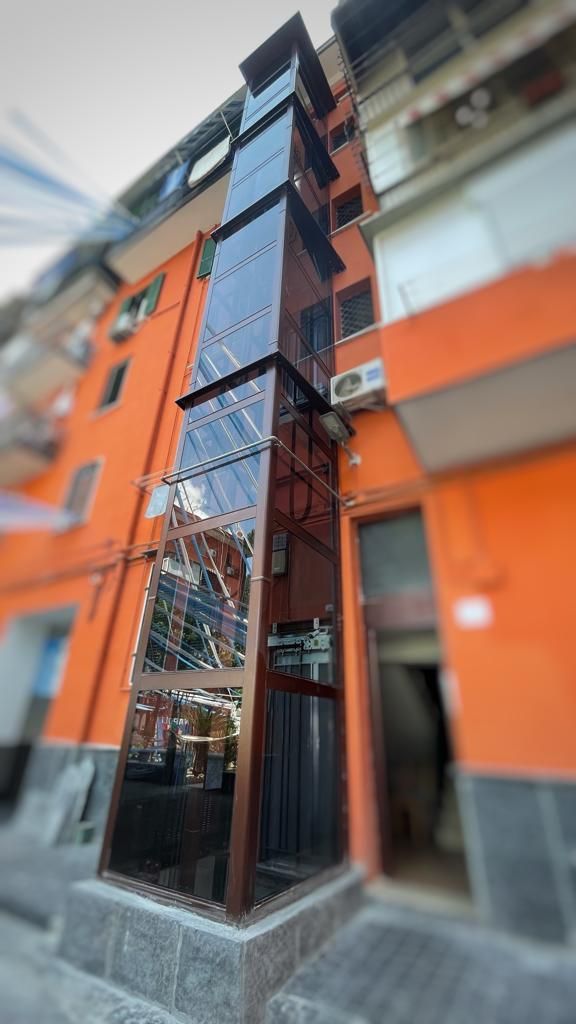 Ascensore in vetro all'esterno di un edificio. Telaio marrone, pannelli trasparenti. Sullo sfondo l'edificio è arancione e grigio.