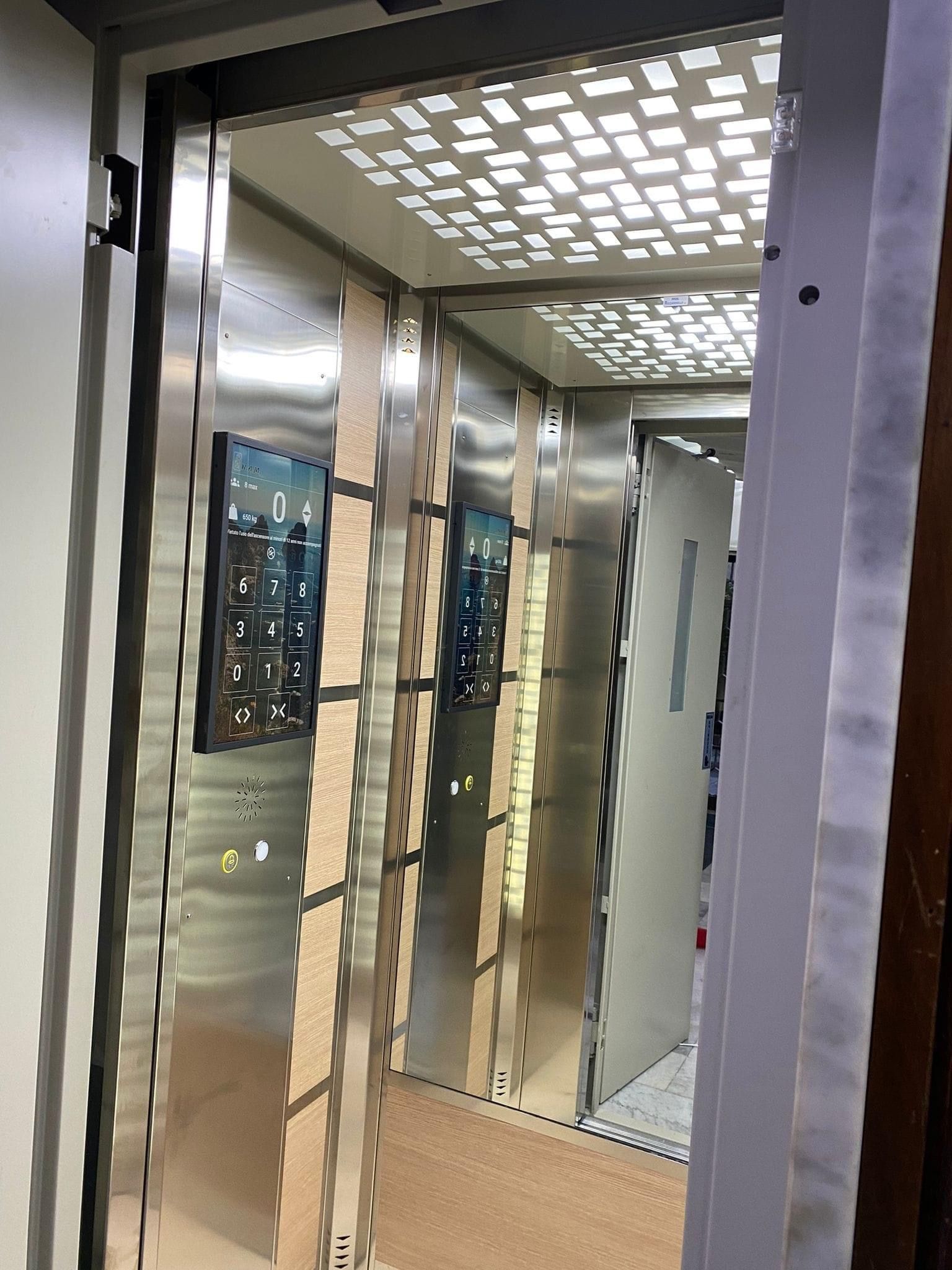 Interno di un ascensore in metallo lucido con soffitto illuminato. Due pannelli di controllo sono visibili su una parete laterale.