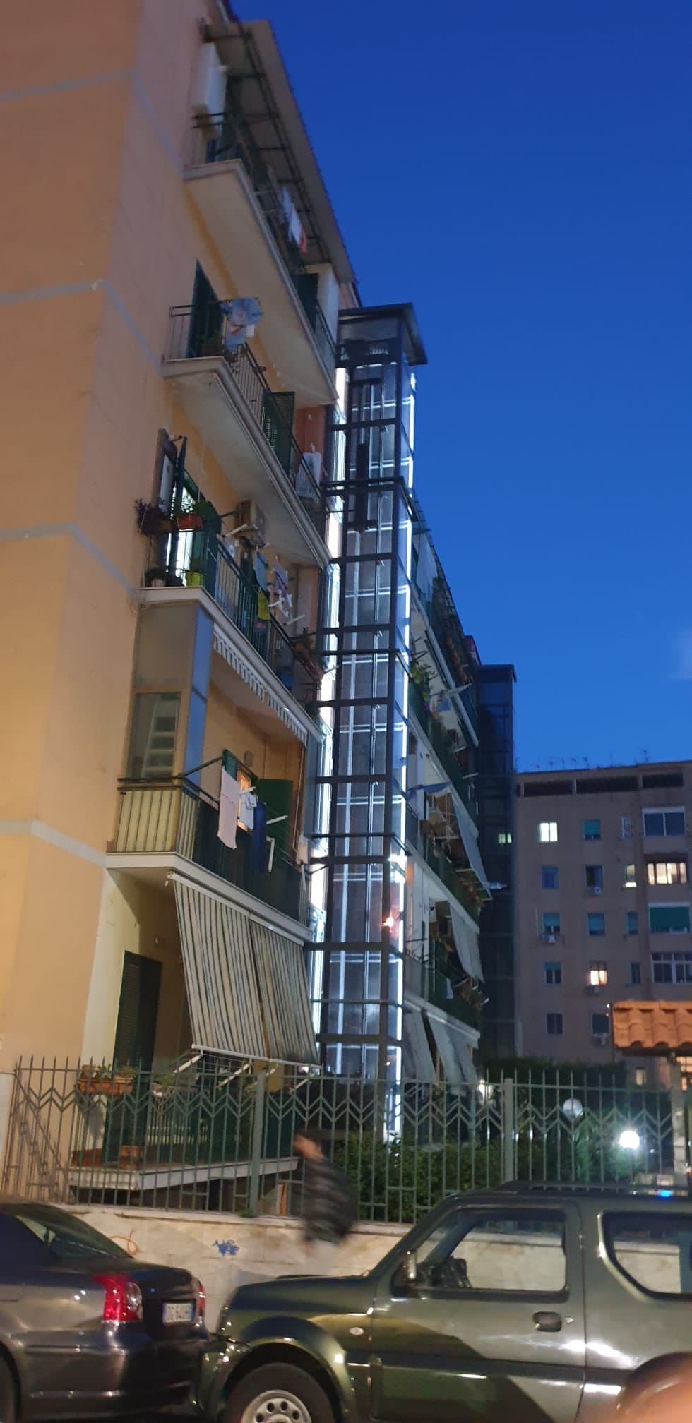 Un alto ascensore in vetro aggiunto all'esterno di un condominio, al tramonto, con auto parcheggiate nelle vicinanze.