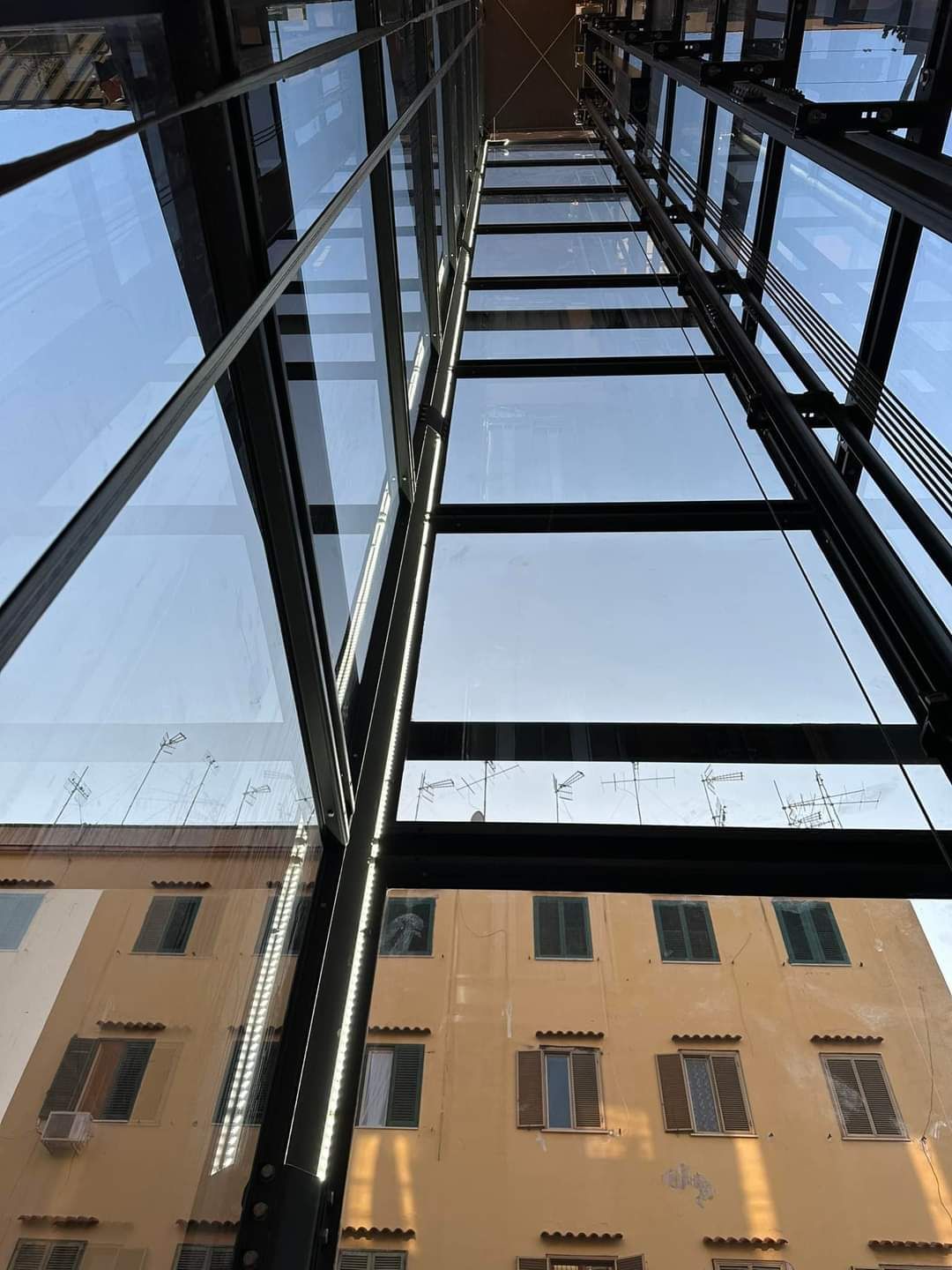 Vano ascensore in vetro con vista esterna di un edificio giallo con numerose finestre e persiane.