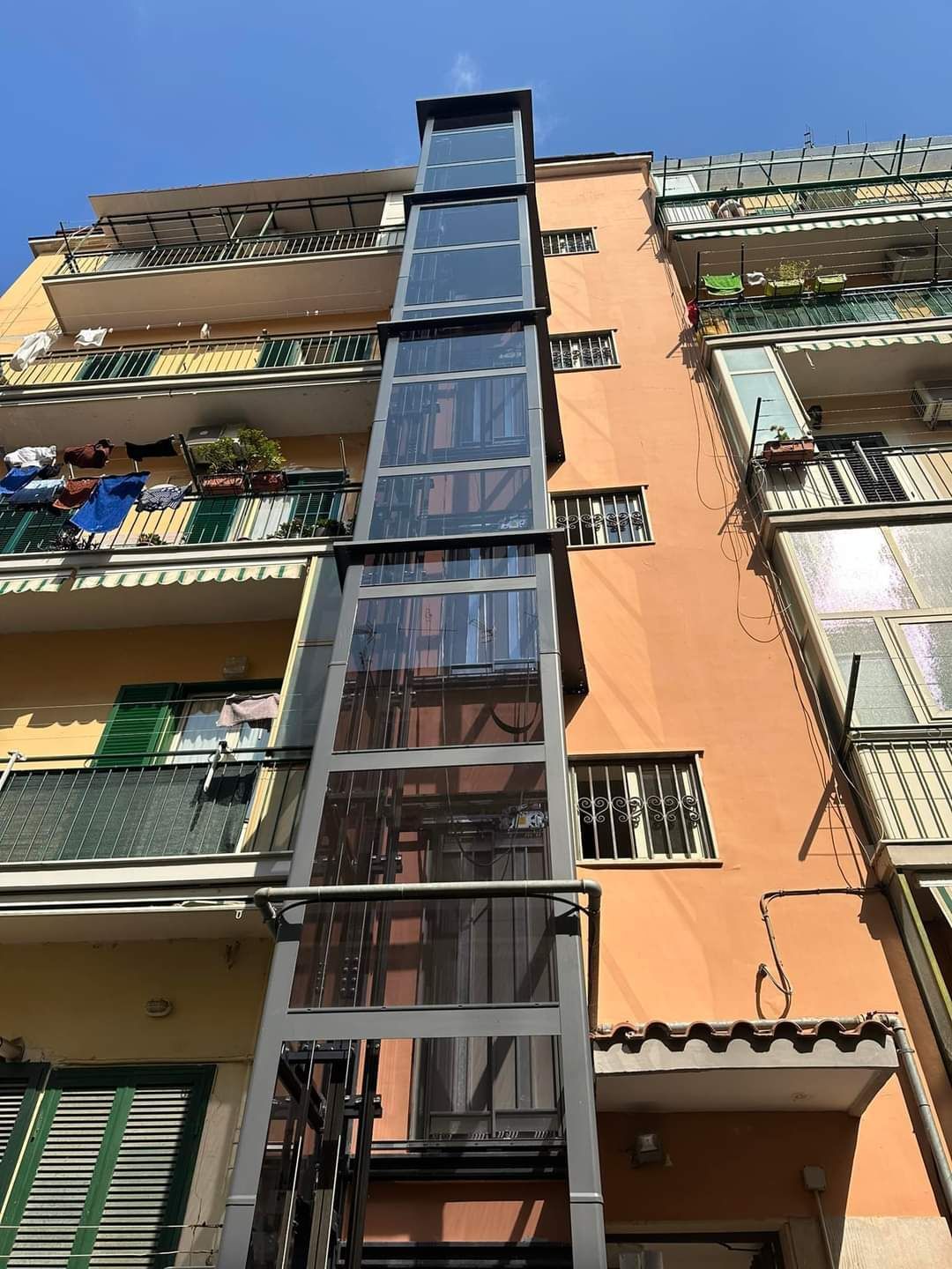 Ascensore esterno in vetro collegato a un edificio a più piani. Struttura in metallo, facciata arancione e gialla.