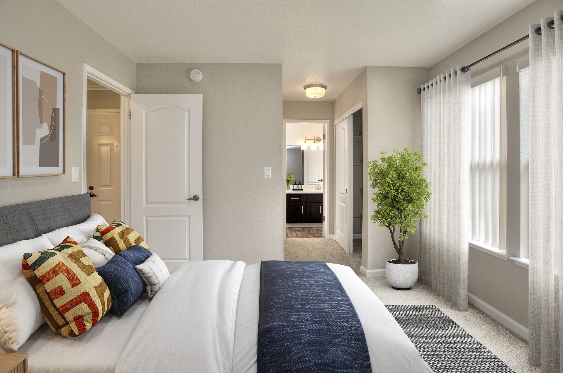 Bedroom with a made bed, decorative pillows, a large window with sheer curtains, and a doorway to a bathroom at Seventeen Mile Drive Village apartments in Pacific Grove, CA.