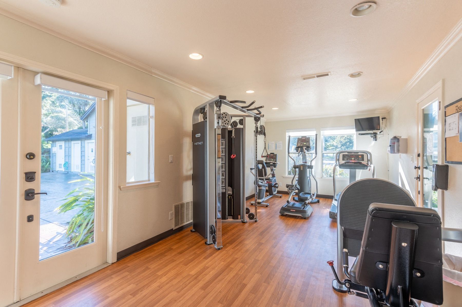 Indoor community gym with cardio equipment and a weight station on wood flooring at Seventeen Mile Drive Village apartments in Pacific Grove, CA.