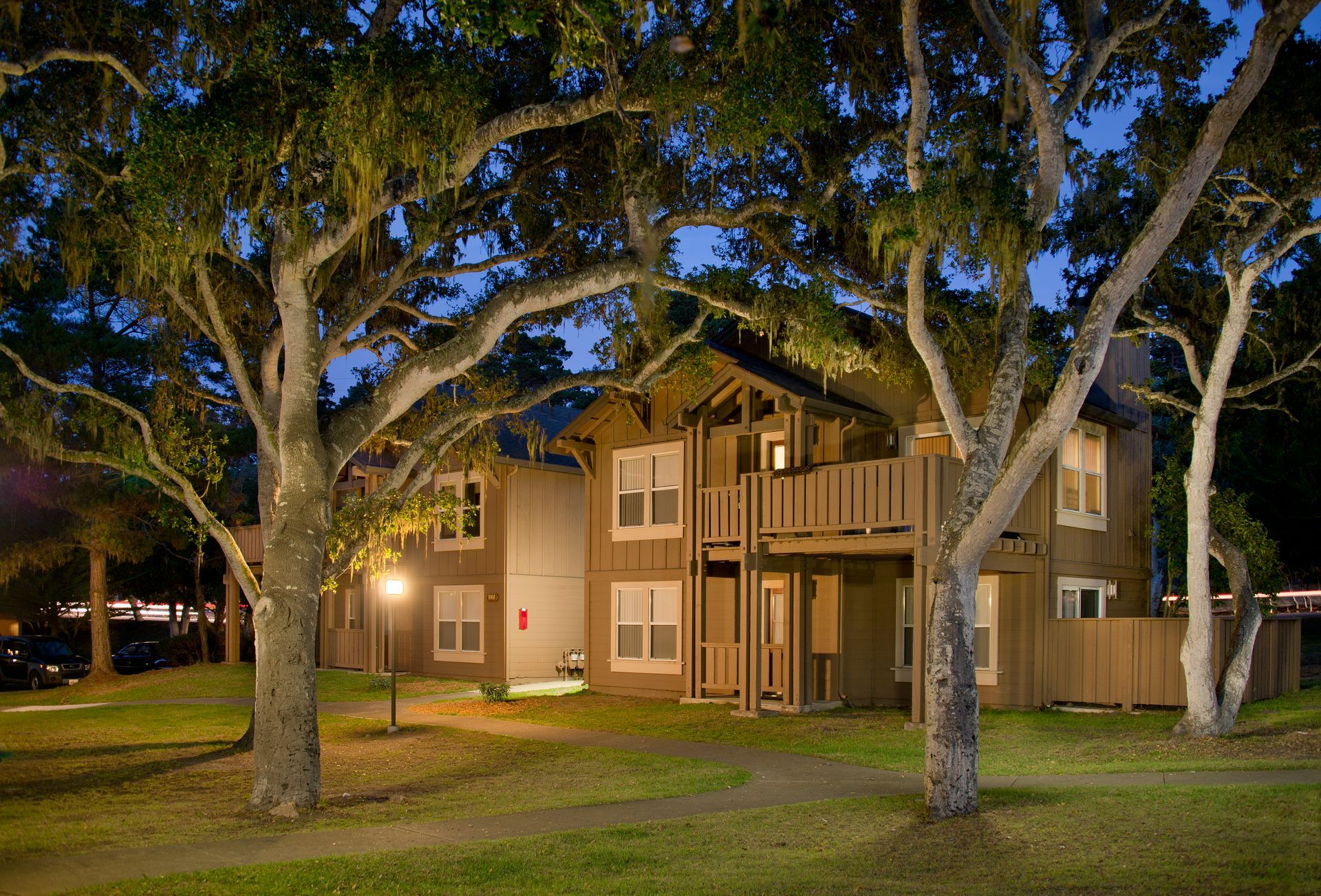 Night view of a two-story apartment building with lit windows and large trees in front at Seventeen Mile Drive Village apartments in Pacific Grove, CA.