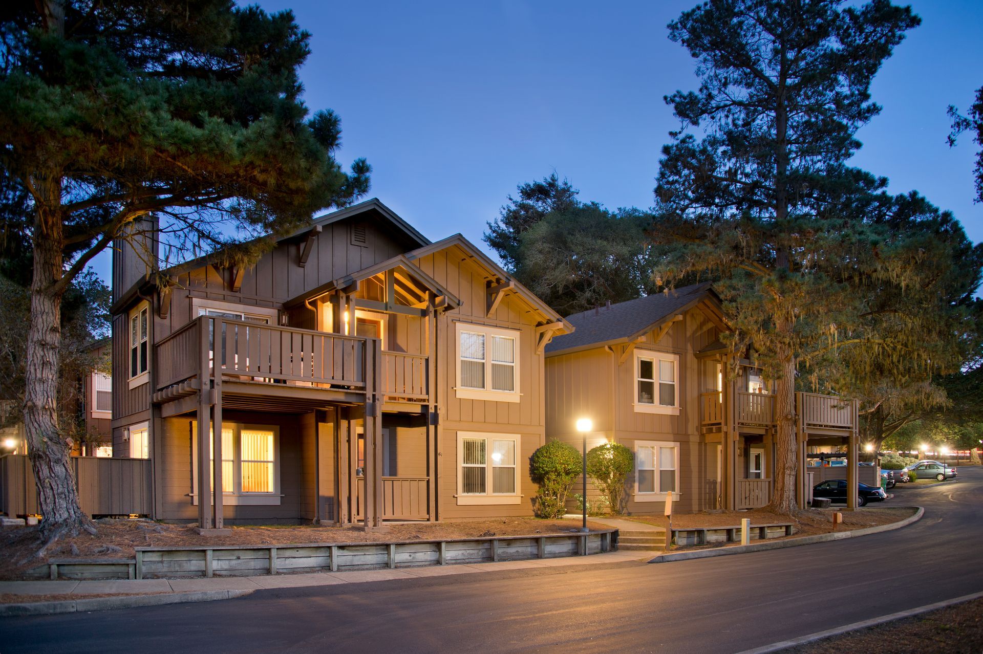 Two-story lodge-style building lit at dusk on a curved road, surrounded by tall pine trees at Seventeen Mile Drive Village apartments in Pacific Grove, CA.