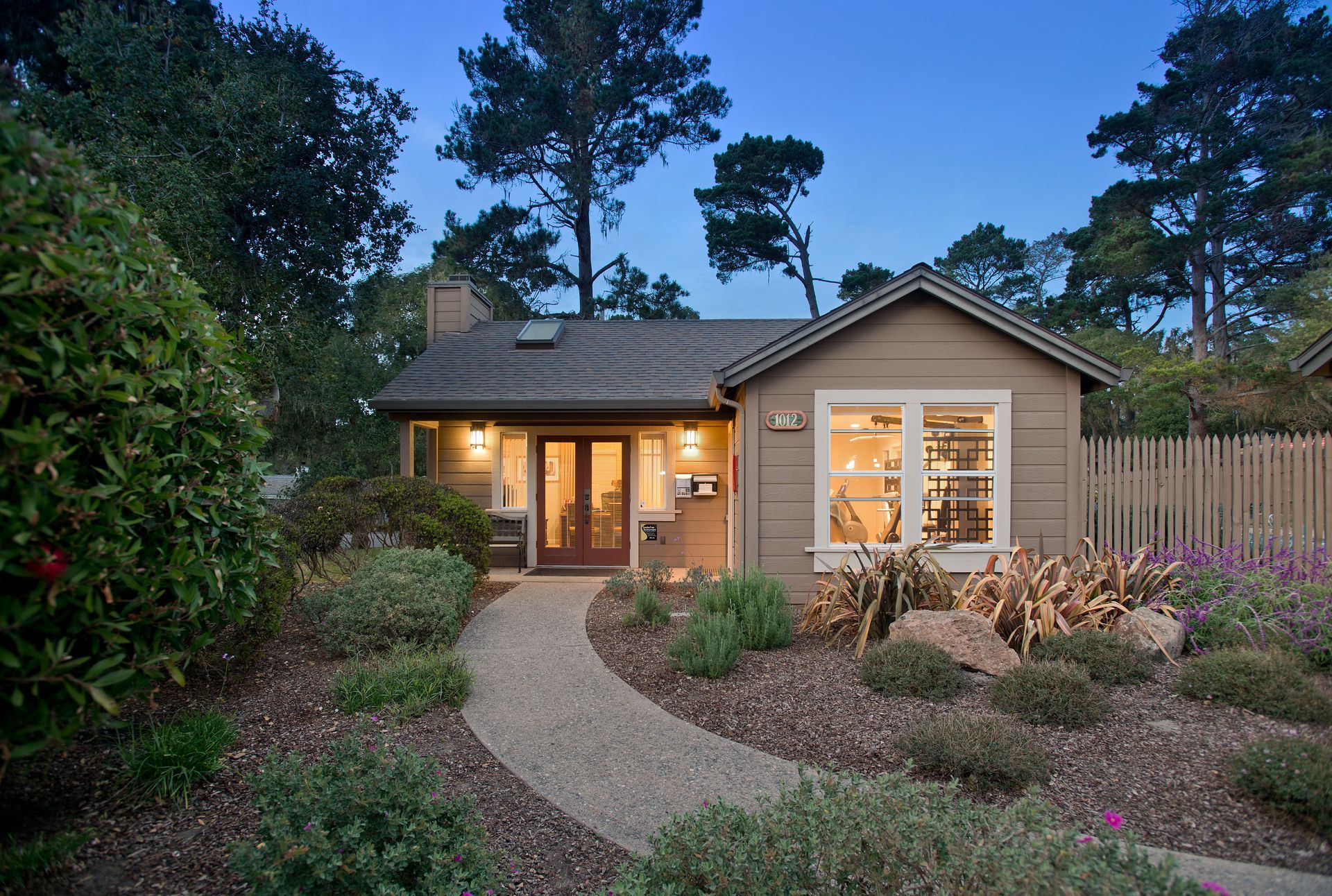 Warmly lit cottage with a curved stone path, garden beds, and trees at dusk at Seventeen Mile Drive Village apartments in Pacific Grove, CA.