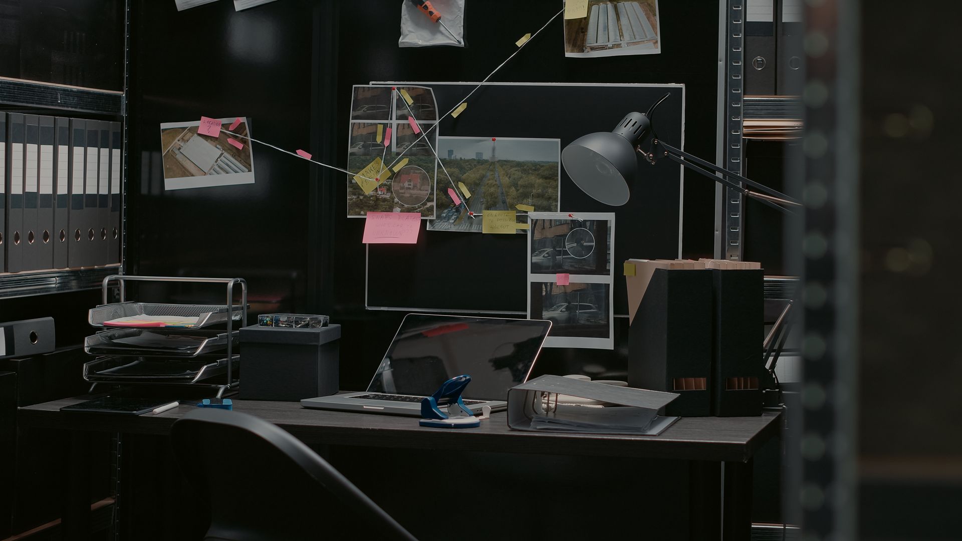A dark office with a desk , chair , computer and a bulletin board.