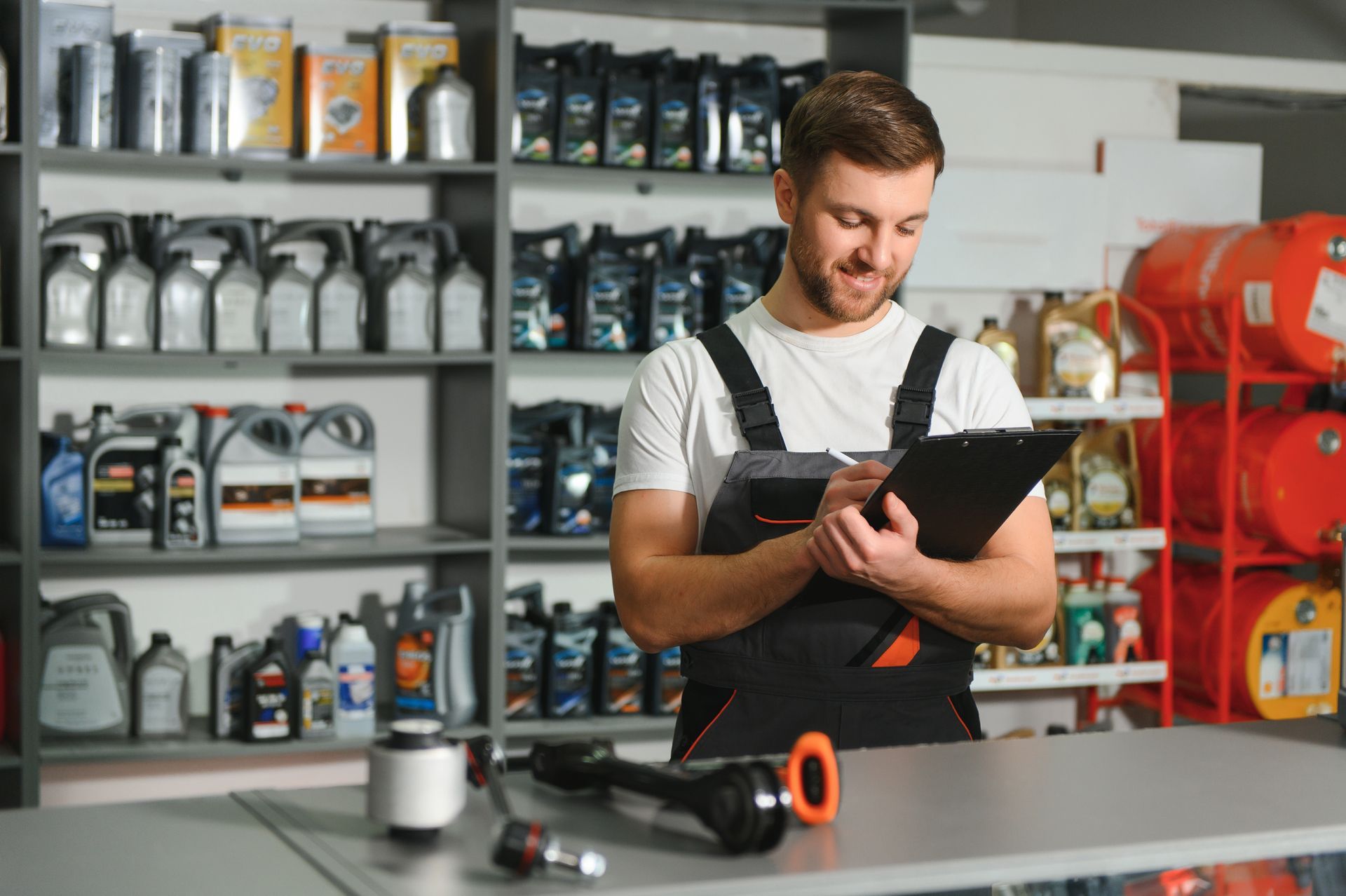 Mechanic writing on clipboard in a shop, surrounded by shelves of oil and car parts.