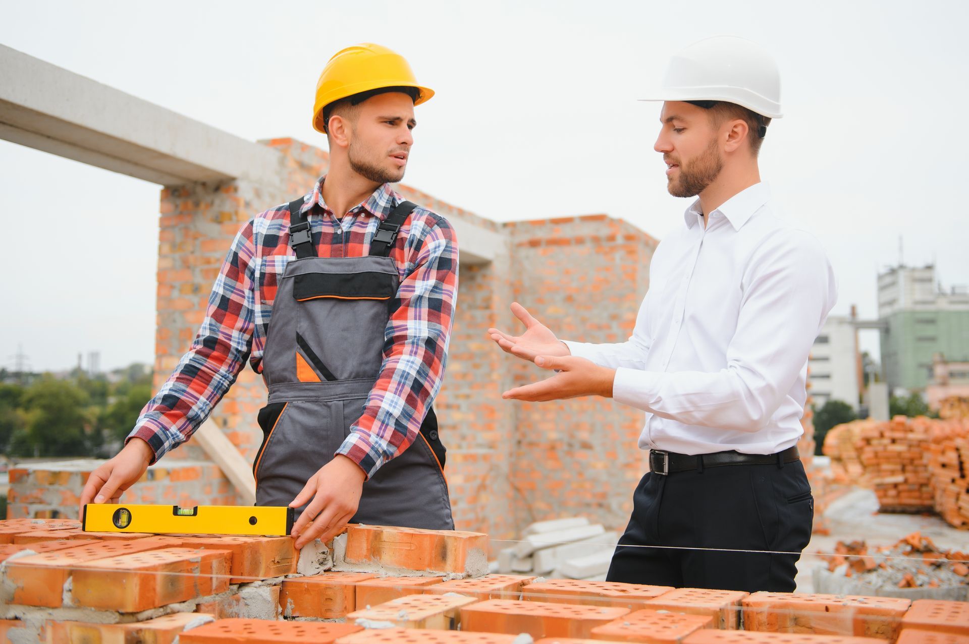 Construction worker using level, discussing with architect at brick construction site.