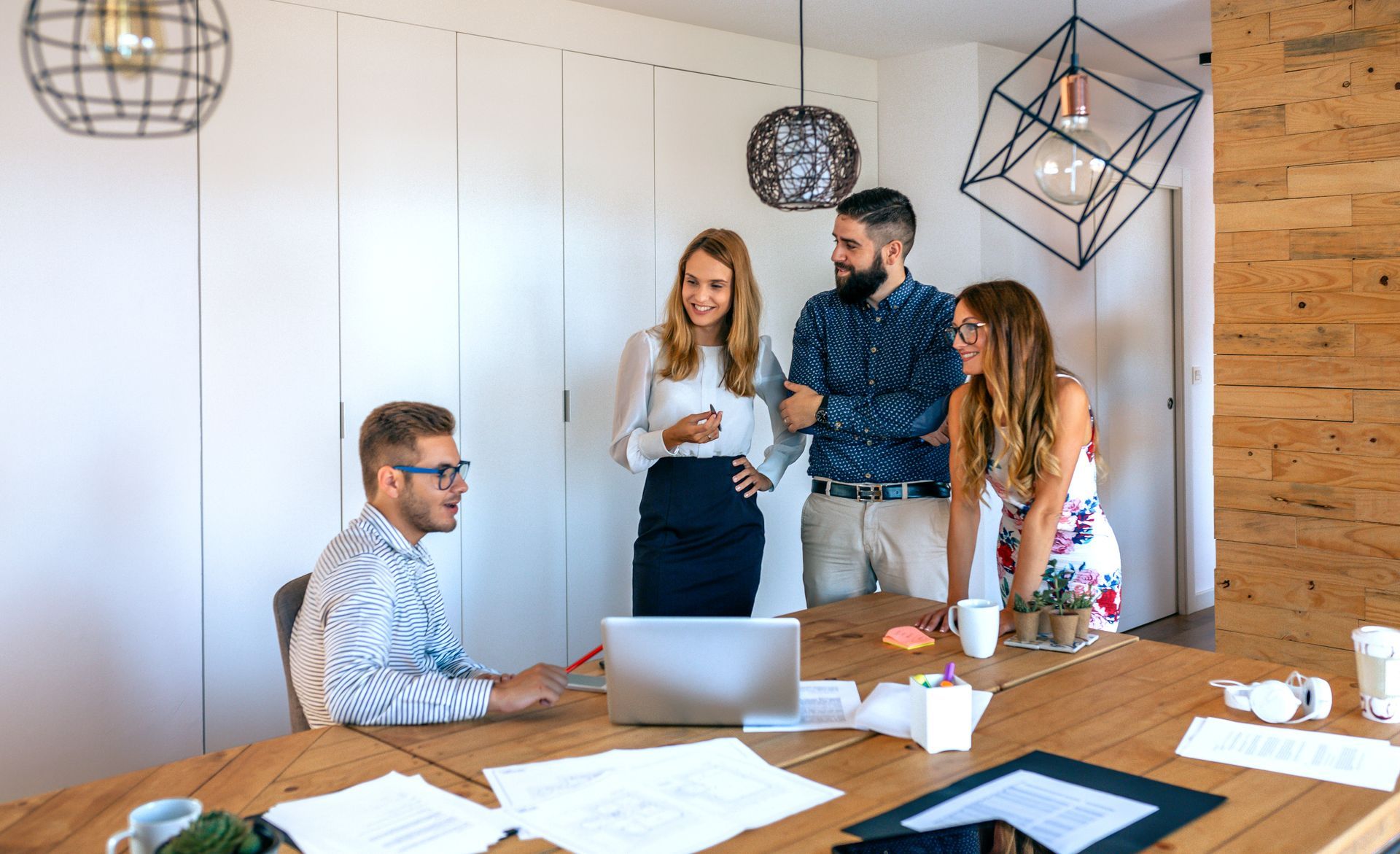 Four office workers collaborating around a large wooden table with a laptop and papers.