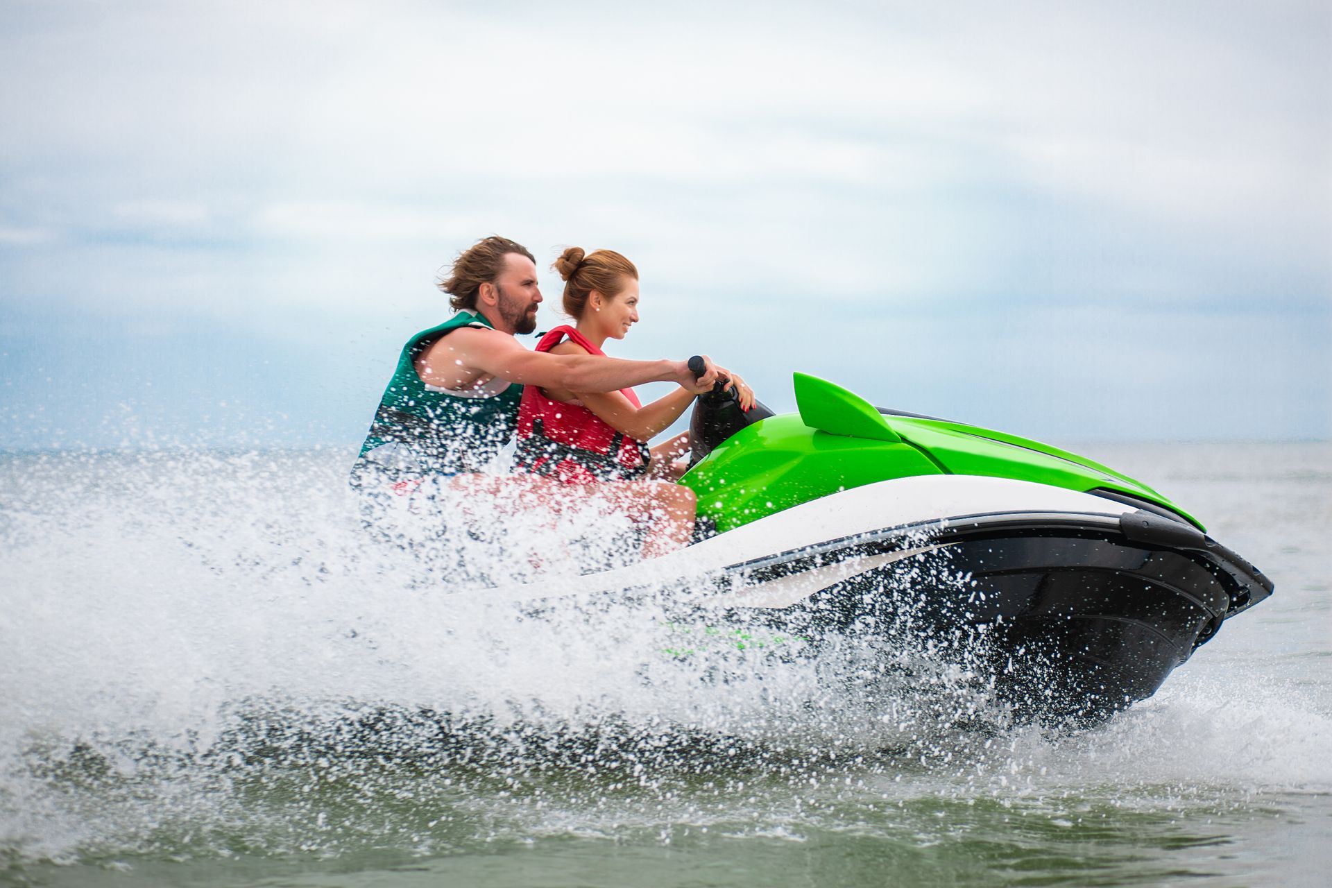Two people ride a green and black jet ski on water, creating a splash. Overcast sky.