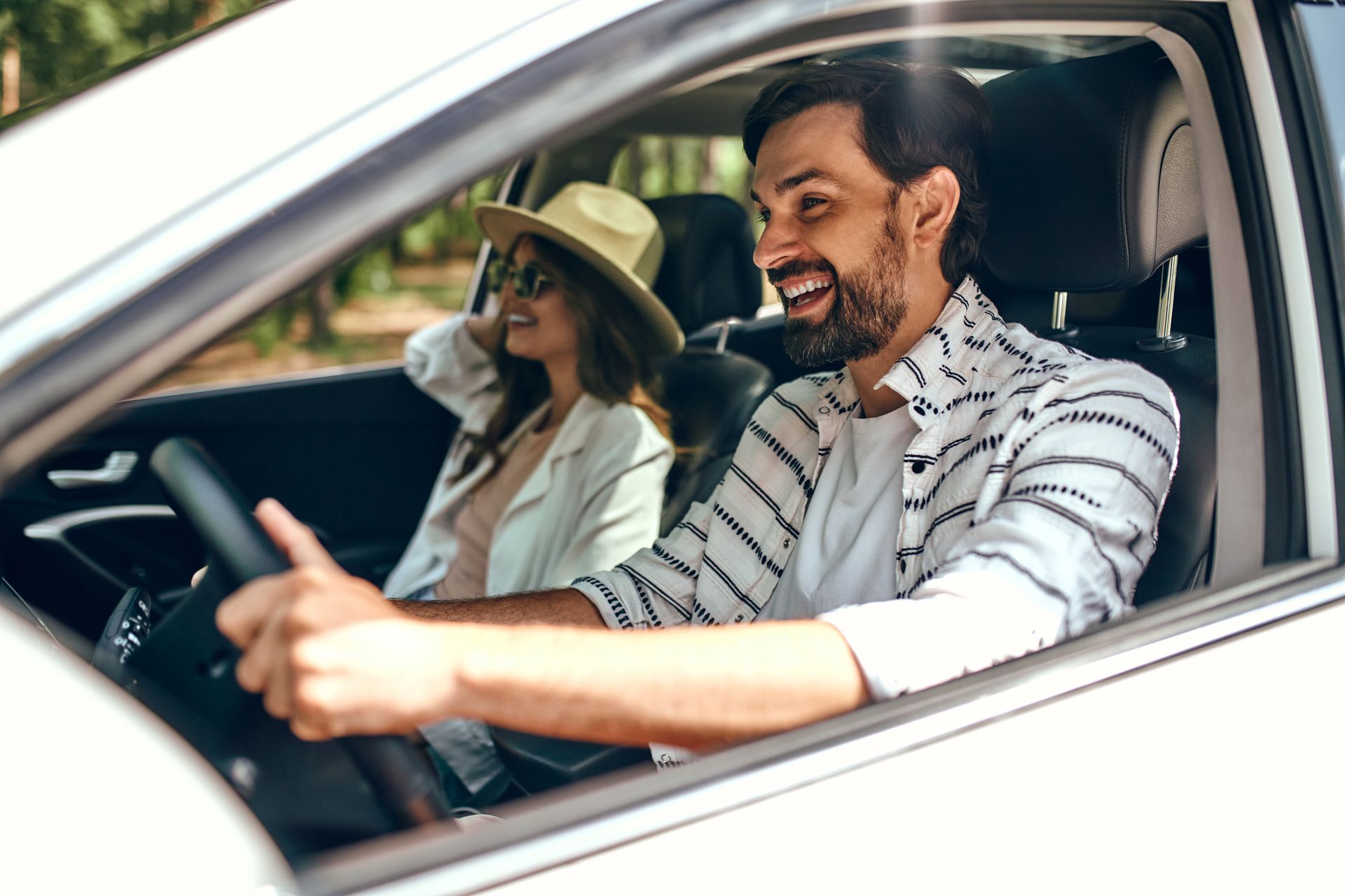 Man smiling while driving a car, a woman in a hat is in the passenger seat.