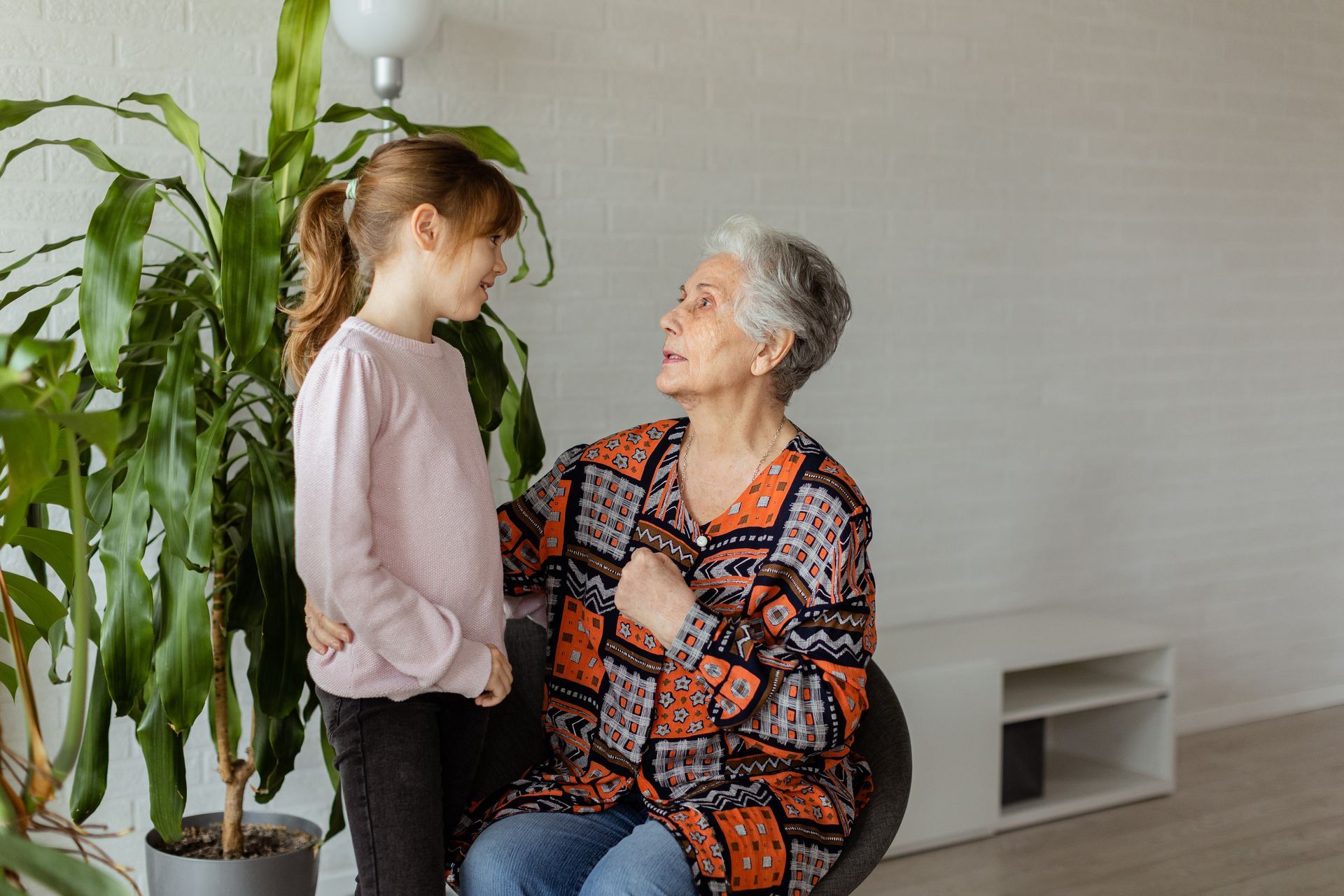Girl talking to woman, woman seated with hand on girl's shoulder, by a plant in room.