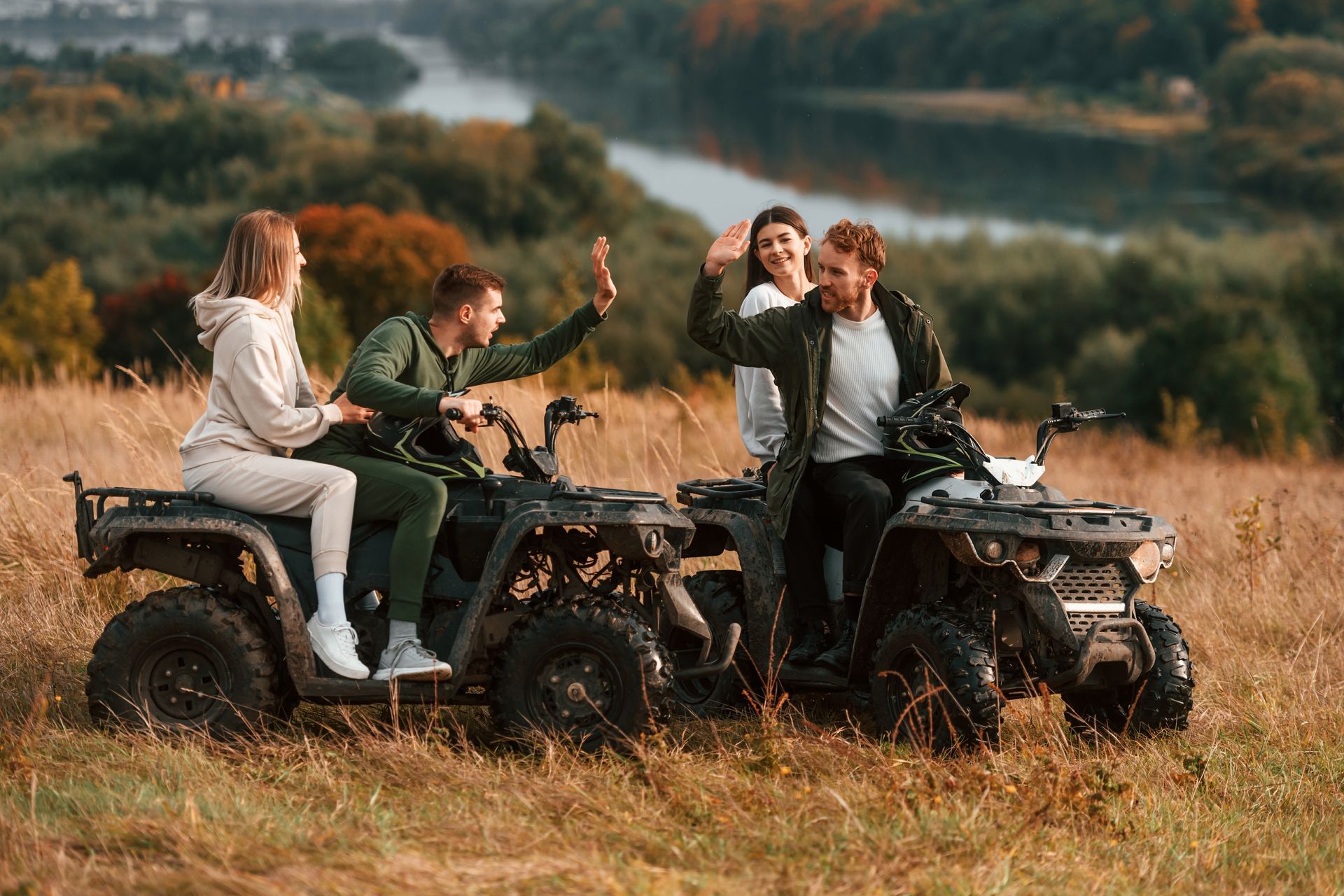 Four people on ATVs in a field, waving at the camera. River and trees in background.