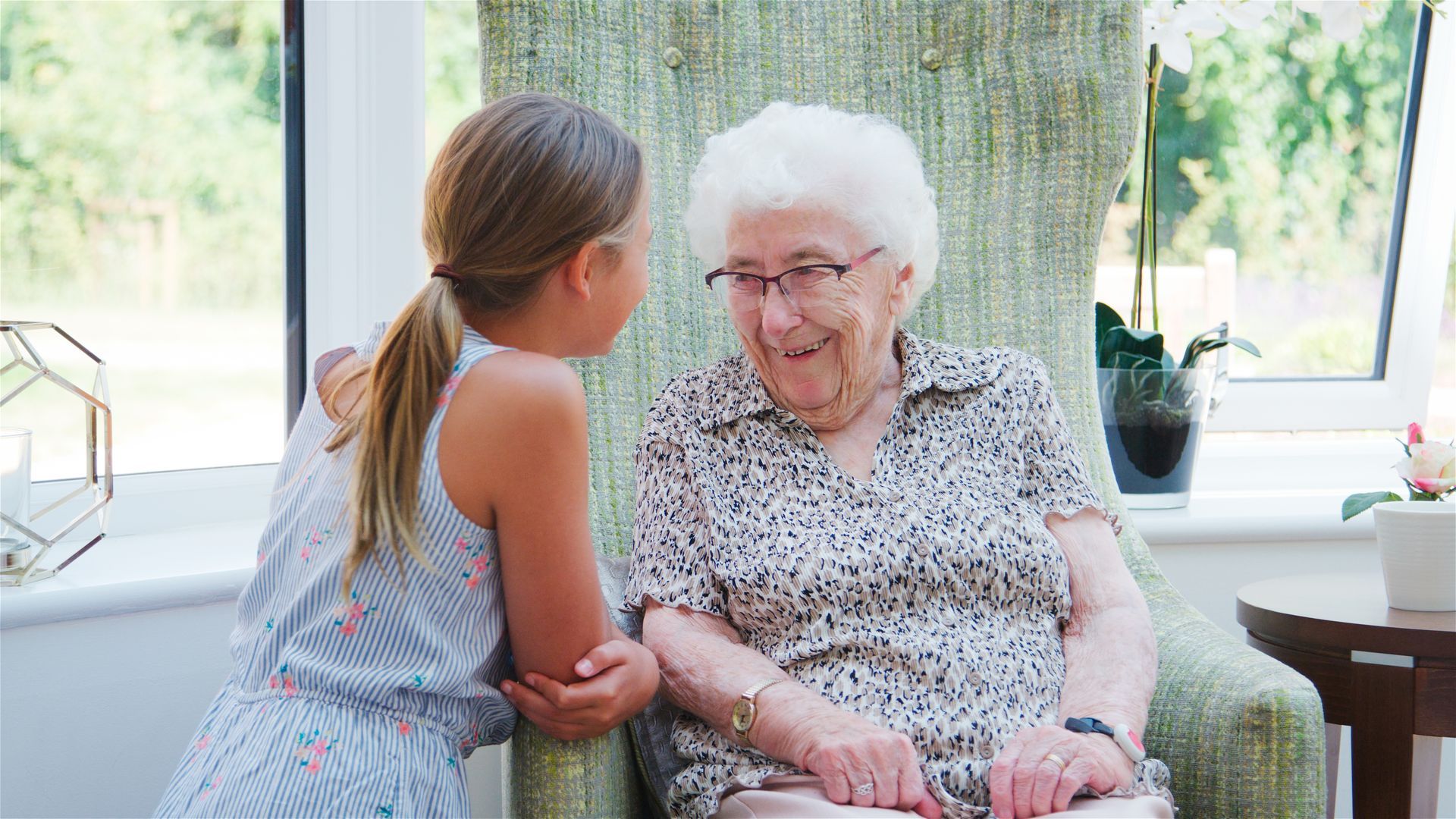 A girl leans toward and talks to an elderly woman in a patterned armchair indoors.