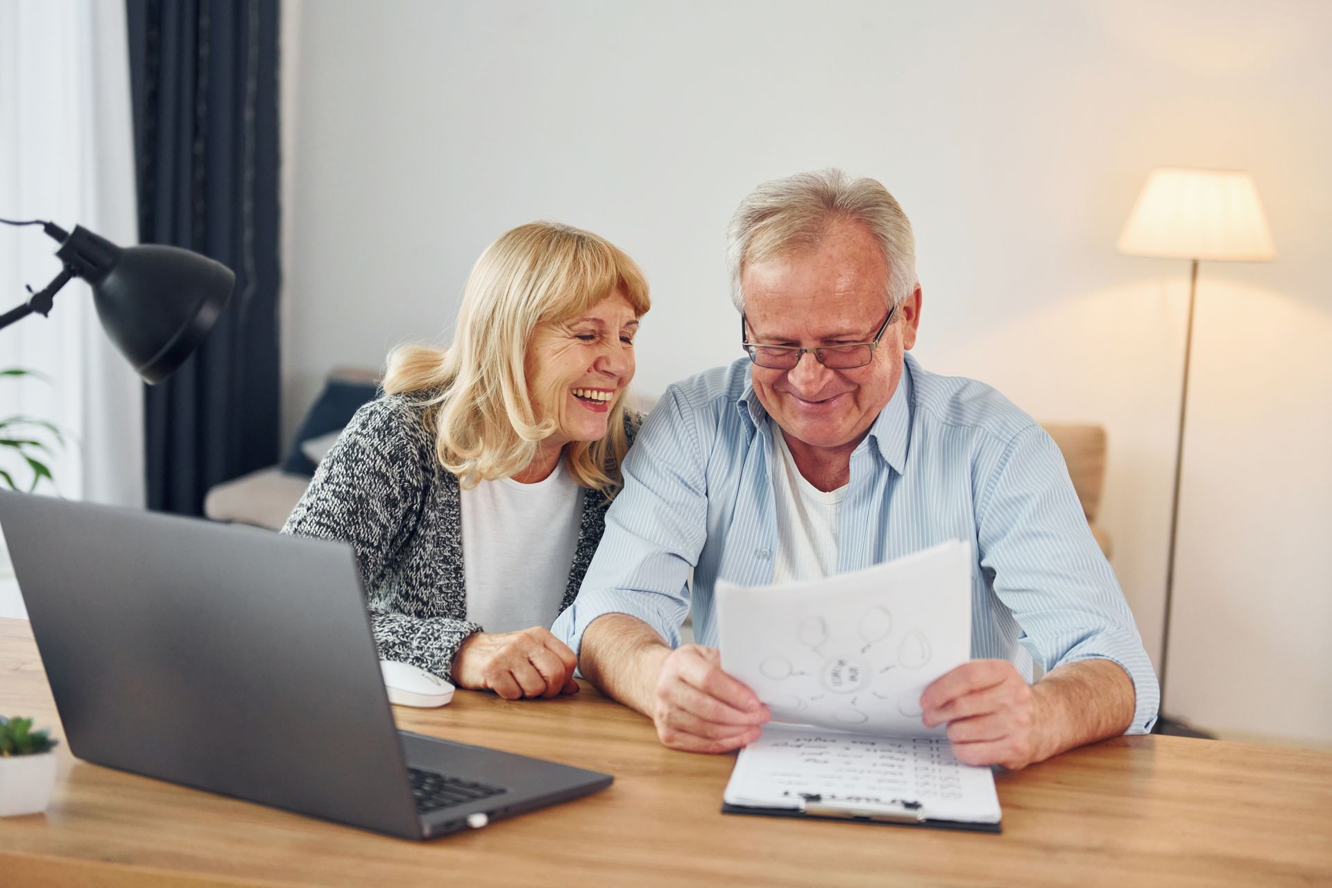 Senior couple looking at papers and laptop at a desk, smiling. Indoor setting.