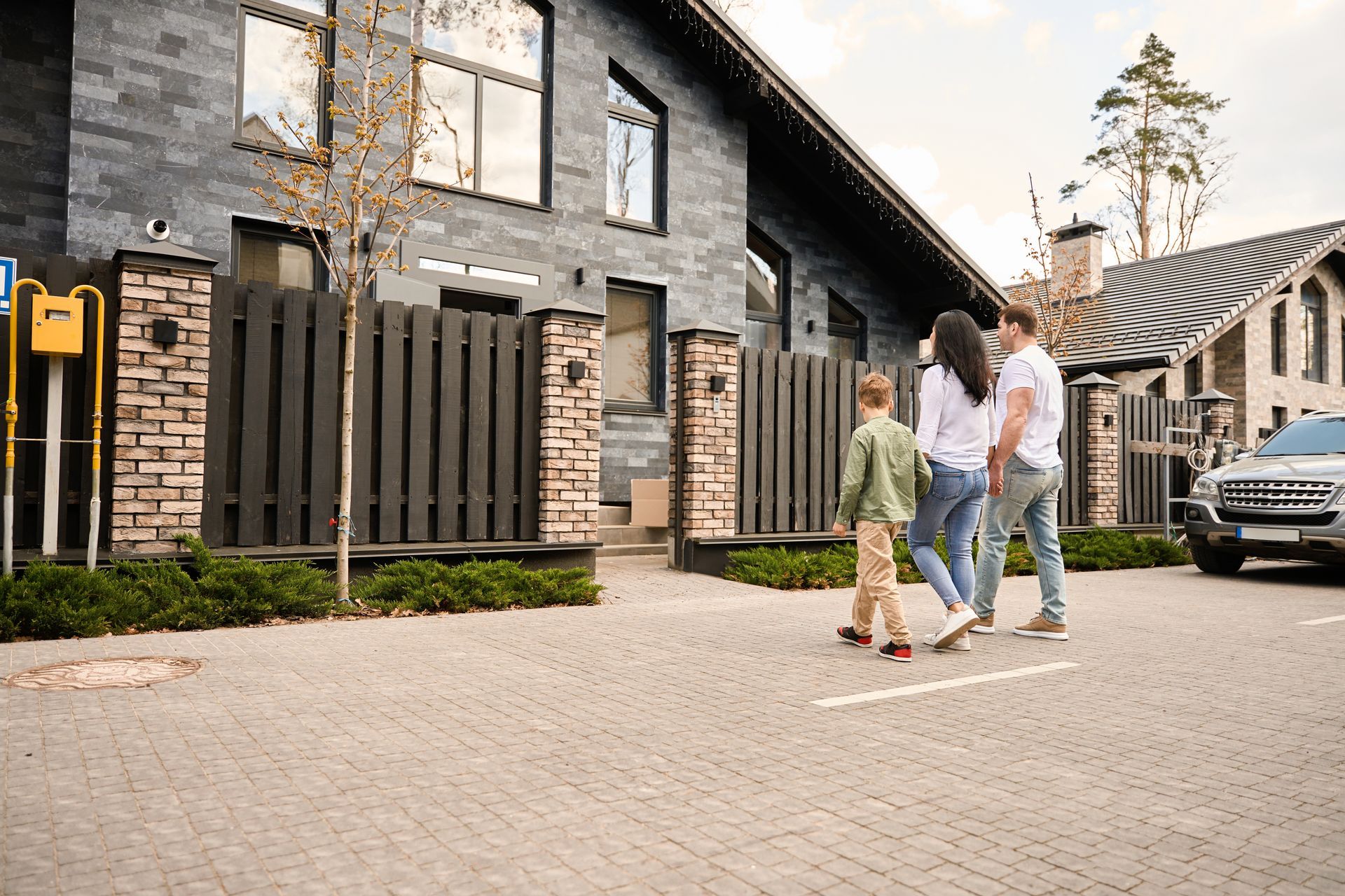 Family walks along a paved street towards a modern home with a dark exterior and fence.