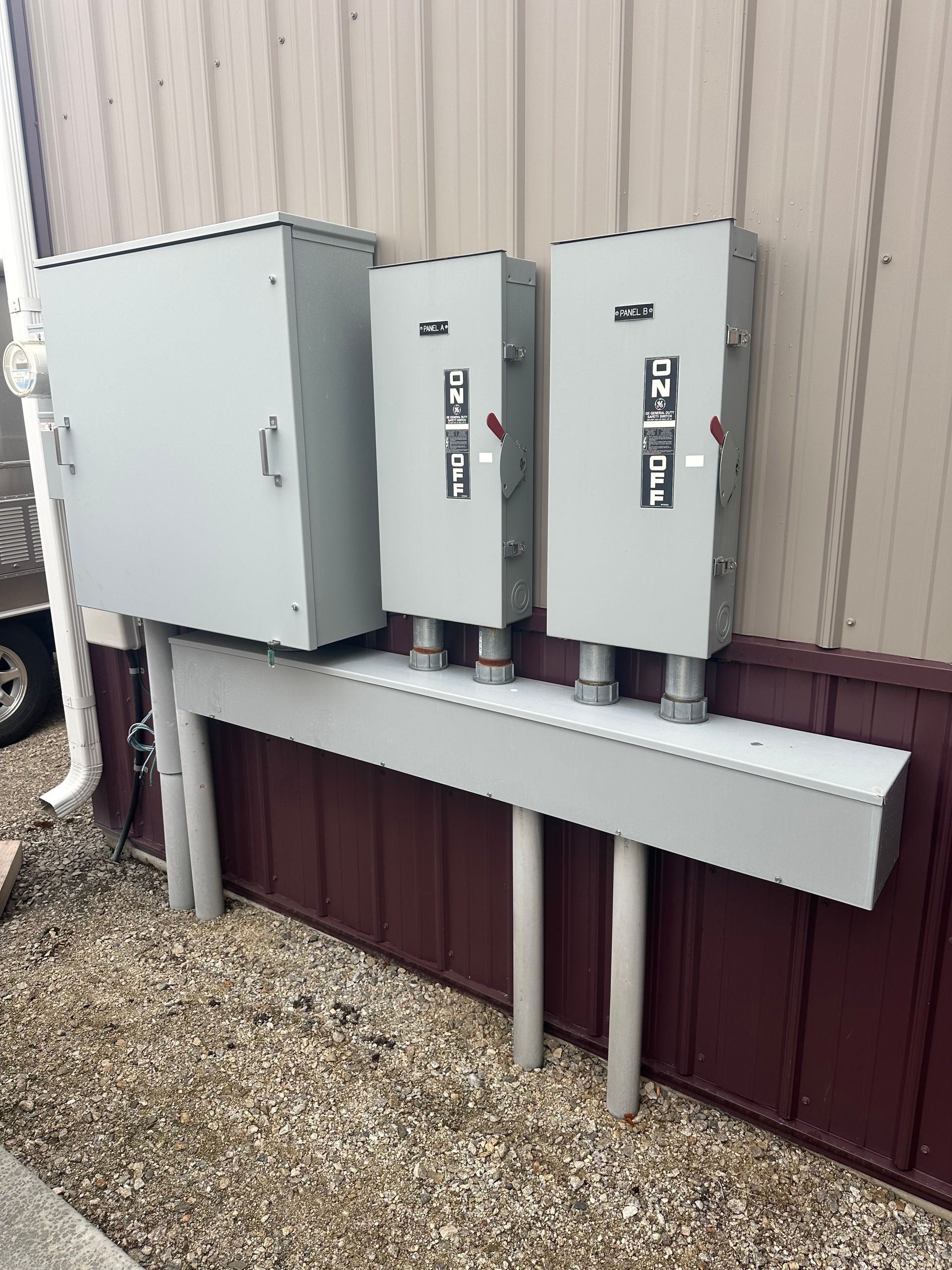 Three gray electrical panels mounted on a beige exterior wall above a maroon foundation.