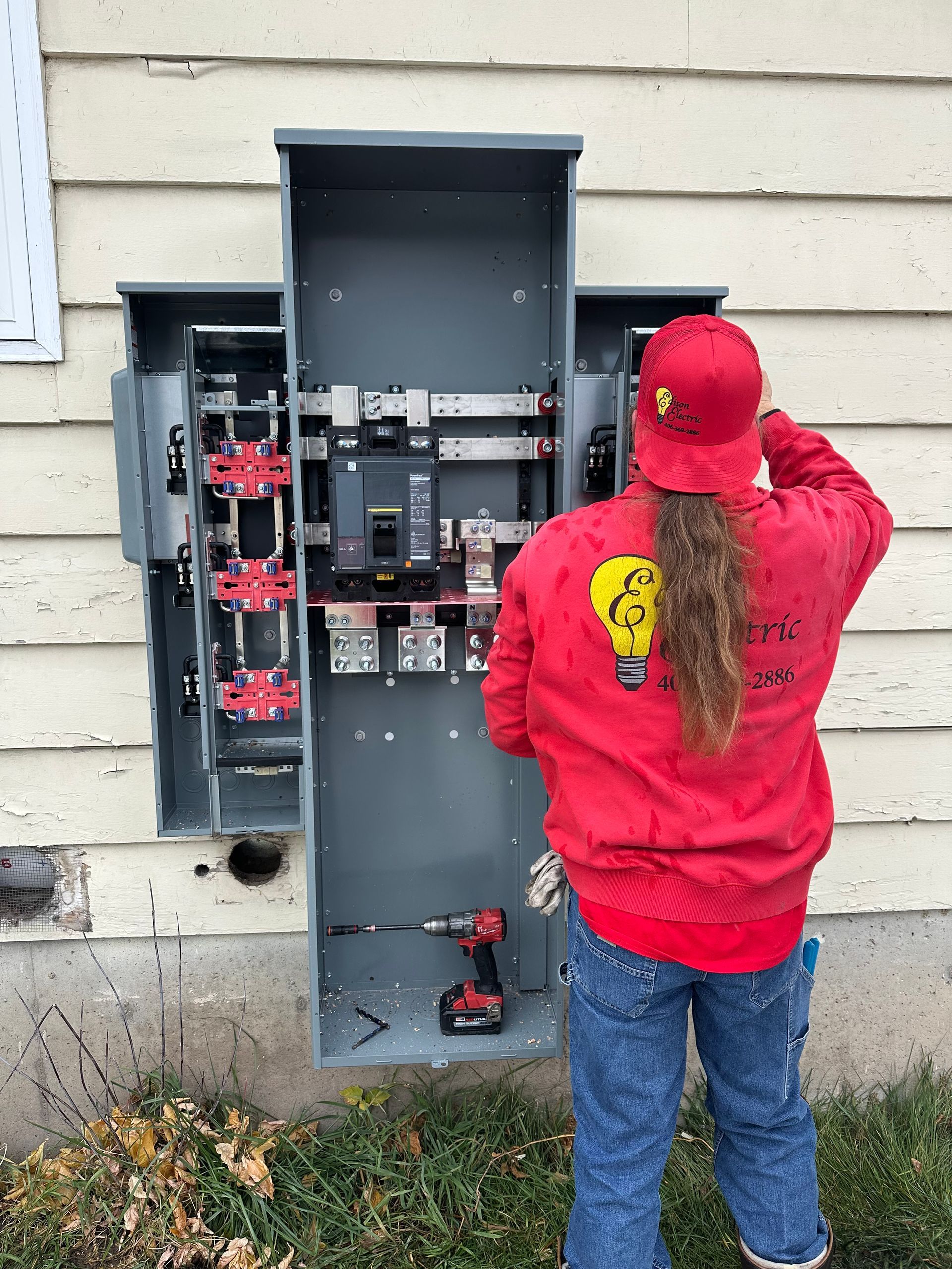 A worker in a red shirt installs an electrical panel on the exterior of a light-colored wood-sided building.