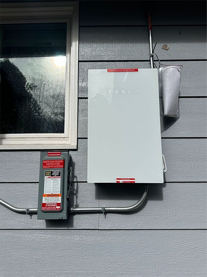 Electric meter and gray breaker box mounted on an exterior wall beside a window.