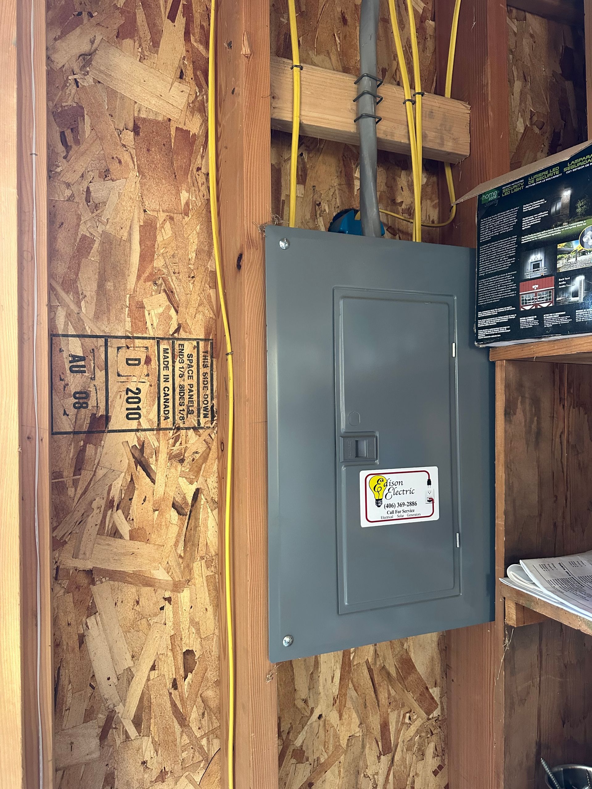 Gray electrical breaker panel on unfinished wooden wall with exposed wiring and pipes.