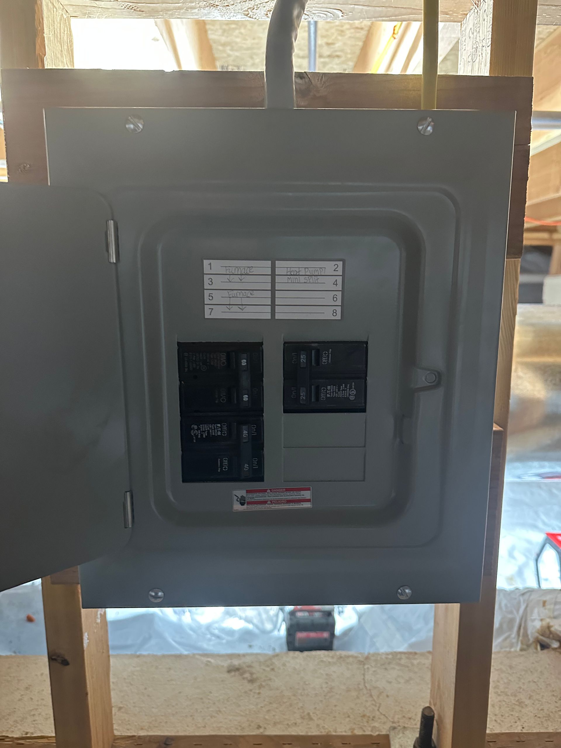 A grey electrical breaker box mounted on unfinished wooden wall studs in a construction setting.