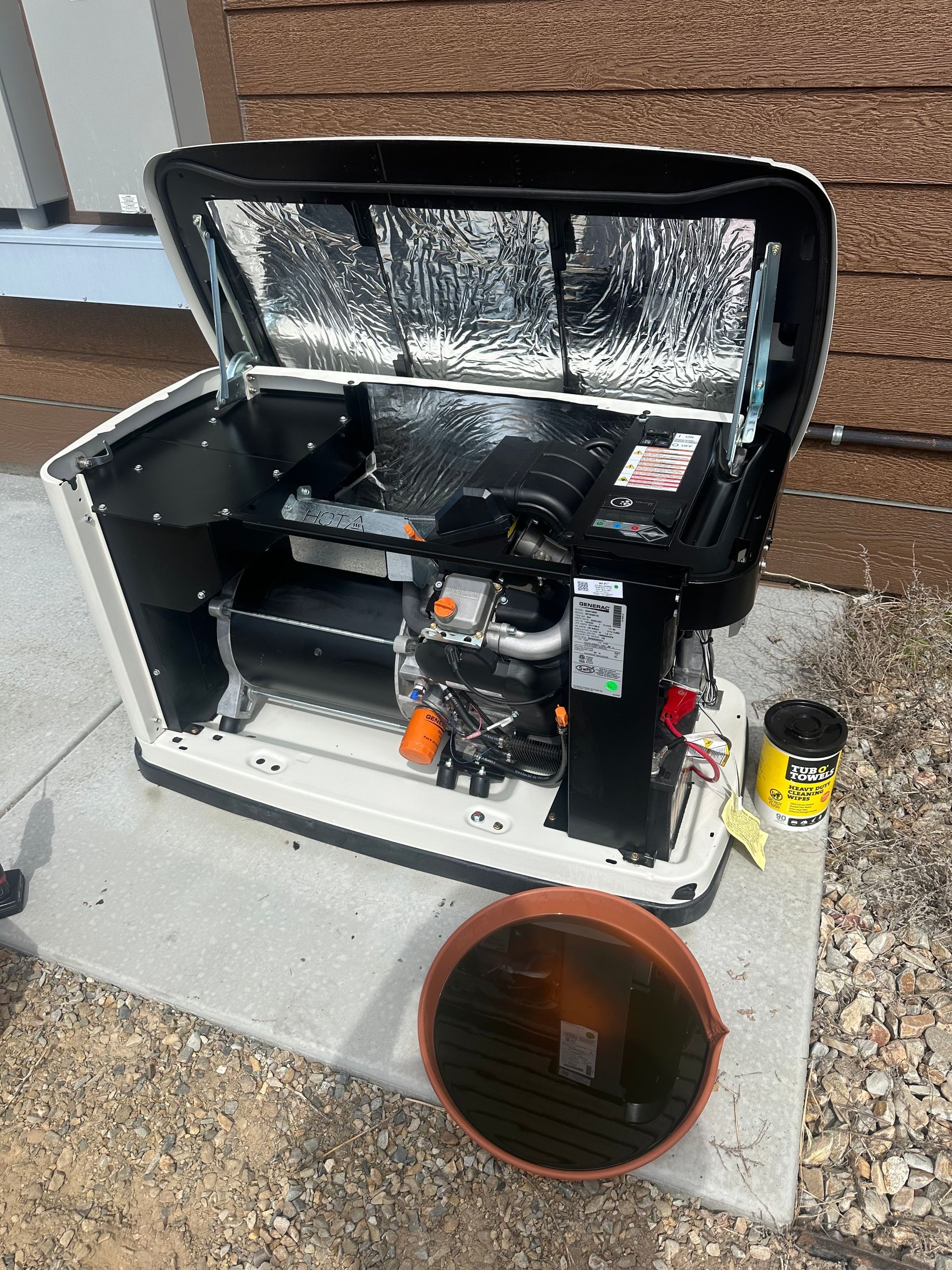 Open HVAC unit with foil-lined lid beside an orange bucket outdoors on a concrete pad.