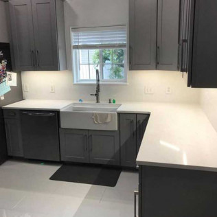 A modern kitchen featuring gray shaker cabinets, white countertops, a white farmhouse sink, and a window above the sink.