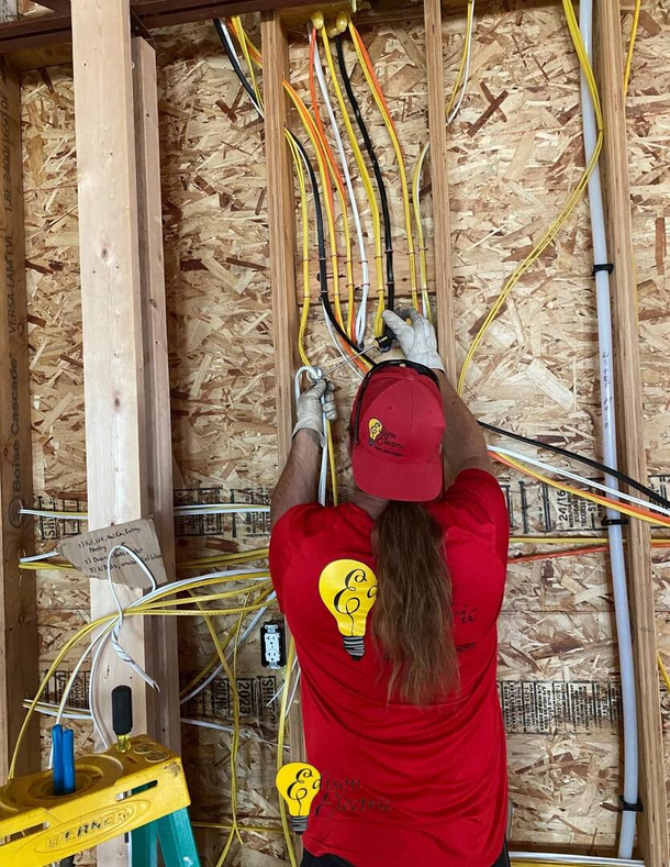 A worker in a red shirt stands between wooden wall studs, installing electrical wiring in a construction site.