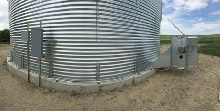 A large, corrugated metal grain bin sits on a concrete foundation in a field, with electrical boxes and a fan unit attached.