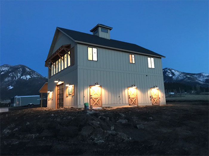 Lit white farmhouse at dusk with warm porch lights, mountains in the background.