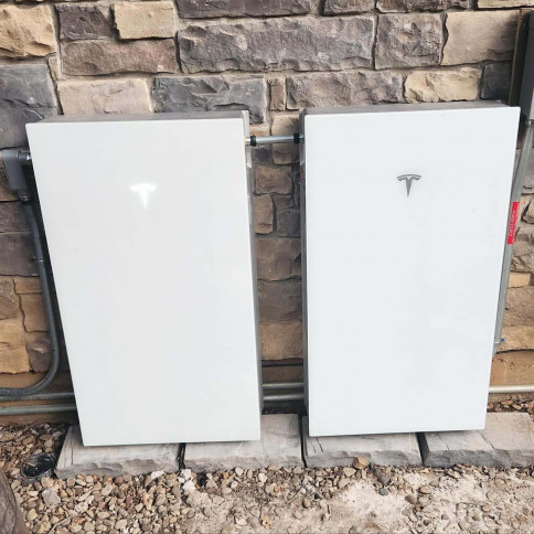 Two white, rectangular Tesla Powerwall home battery units mounted side-by-side on an outdoor stone wall.