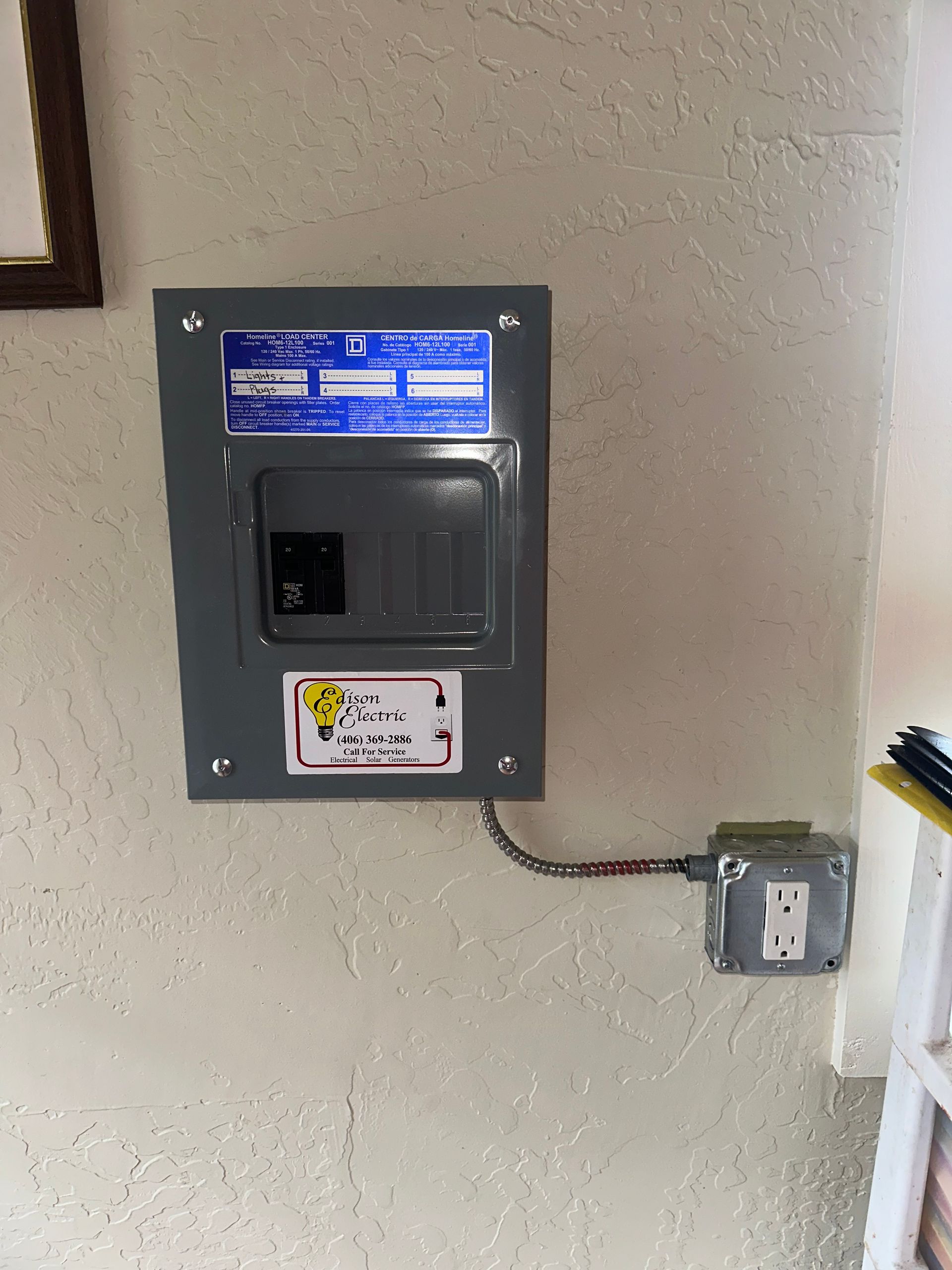 Wall-mounted electrical breaker box with labels and a conduit leading to a small outlet box.