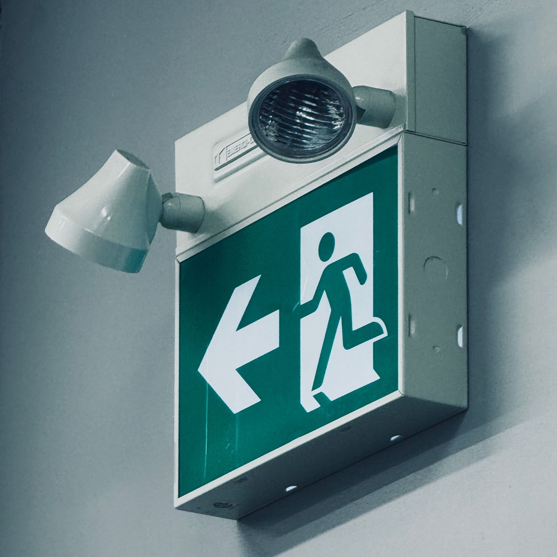 Green illuminated exit sign with a left arrow and running figure on a wall, with two lights mounted above and beside it